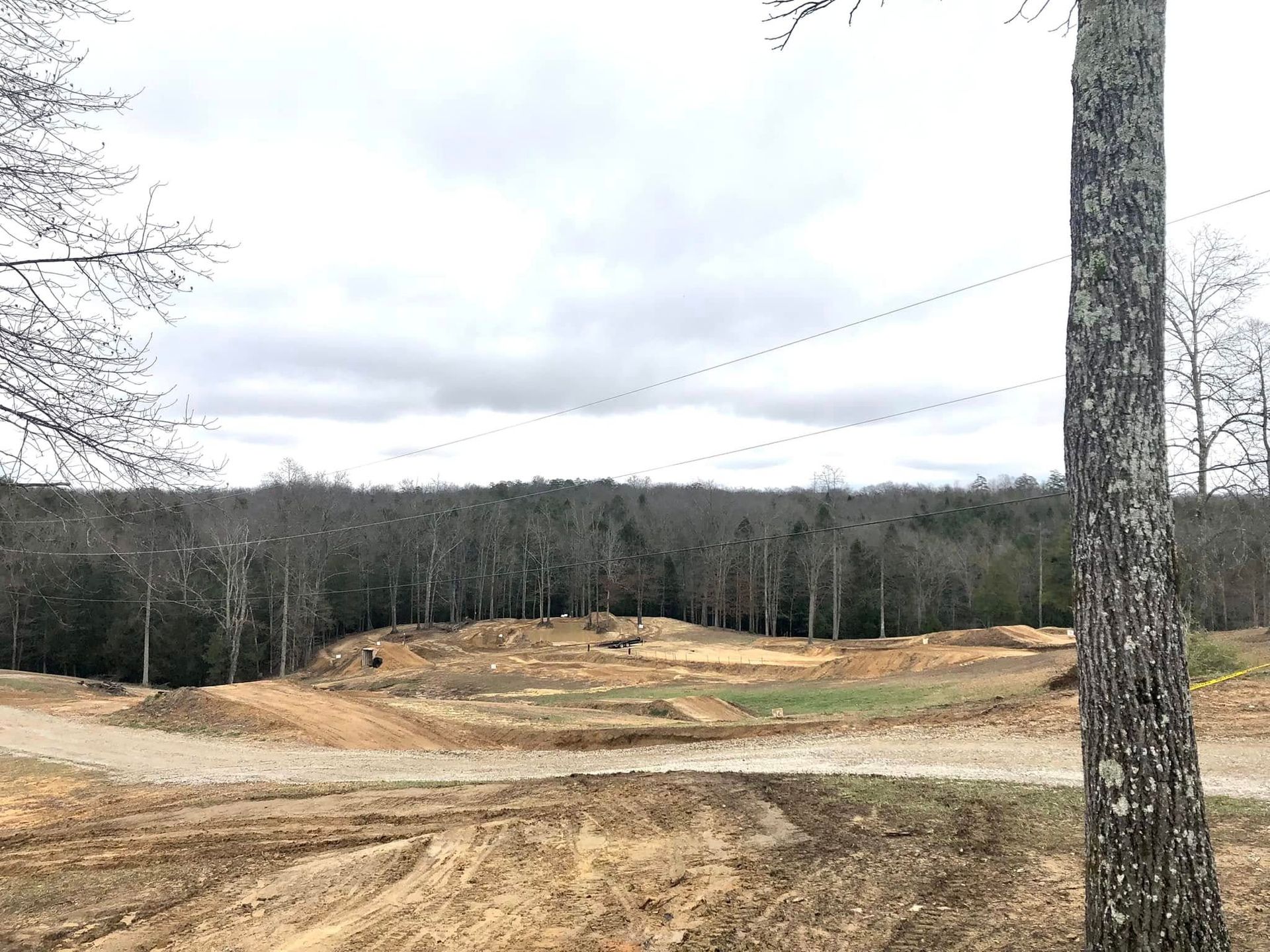 A dirt-covered clearing with mounds and paths in a wooded landscape under a cloudy sky, seen from a slightly elevated view.