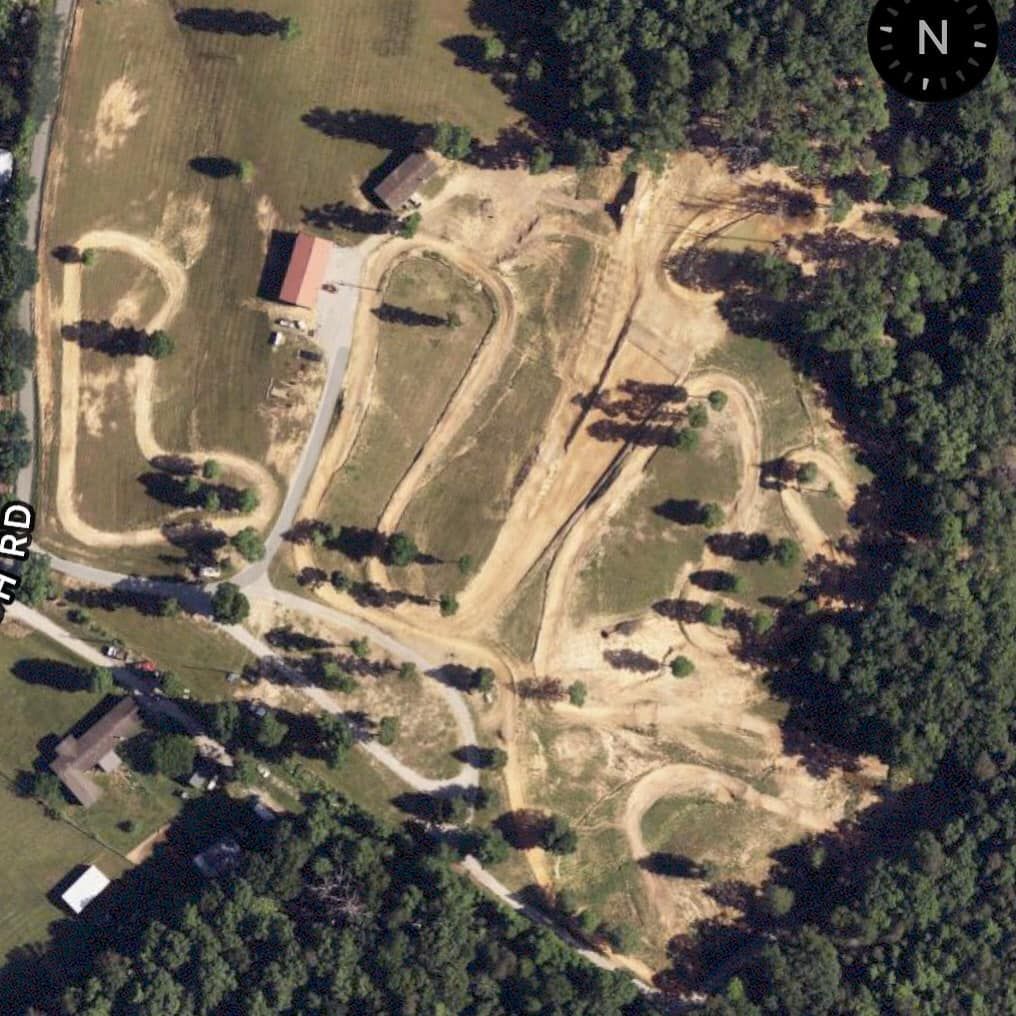 Aerial view of a dirt bike track with winding paths and jumps on a rural property next to buildings and trees.