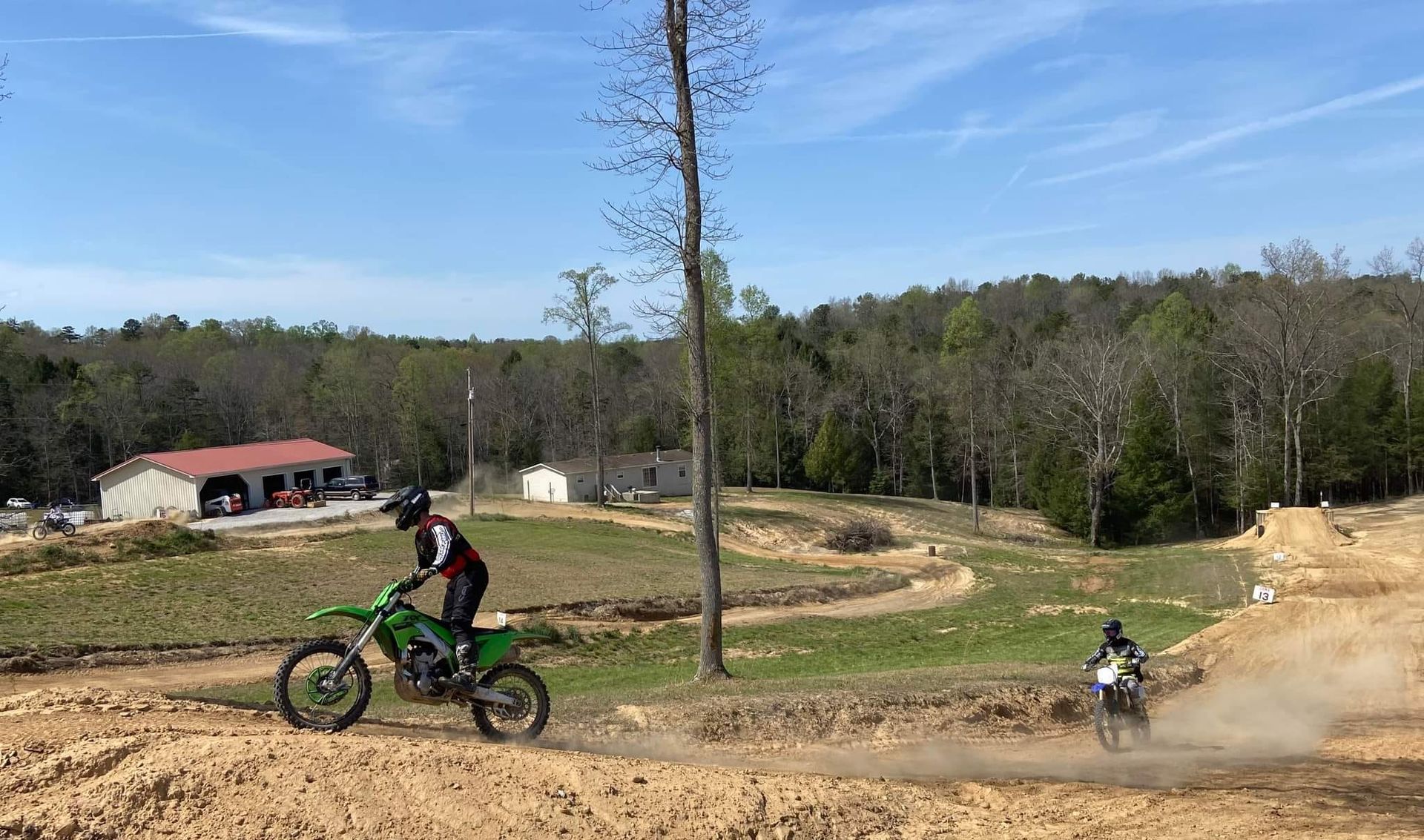 Two dirt bike riders navigate a sandy outdoor track near trees and buildings under a clear blue sky.