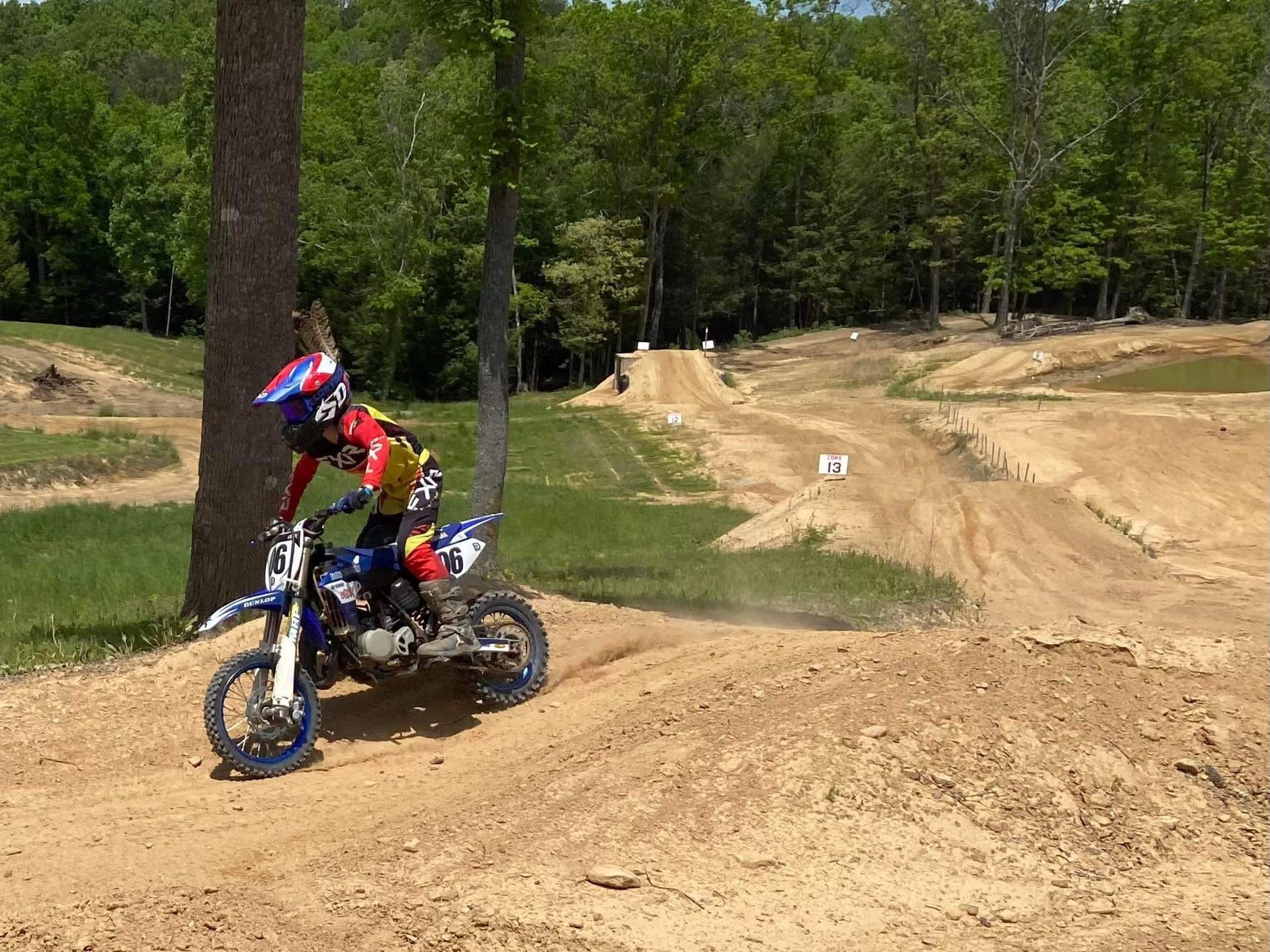 A rider in full gear on a blue dirt bike navigates a sandy track bordered by green trees.