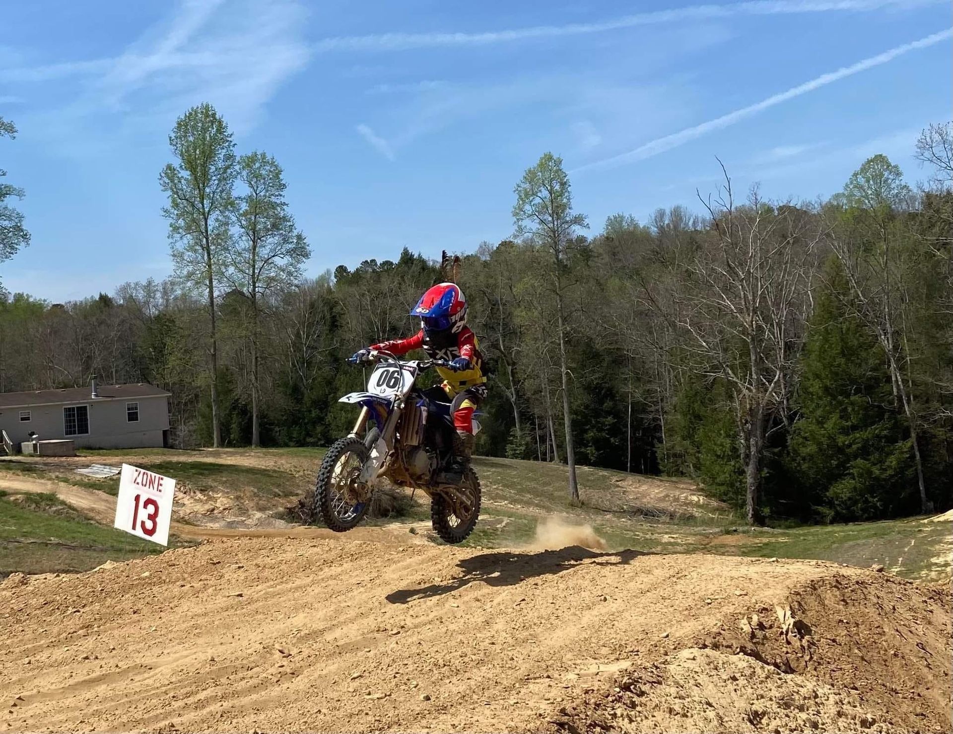 A child in protective gear jumps a dirt bike on a sunny, wooded outdoor track next to a sign marked 