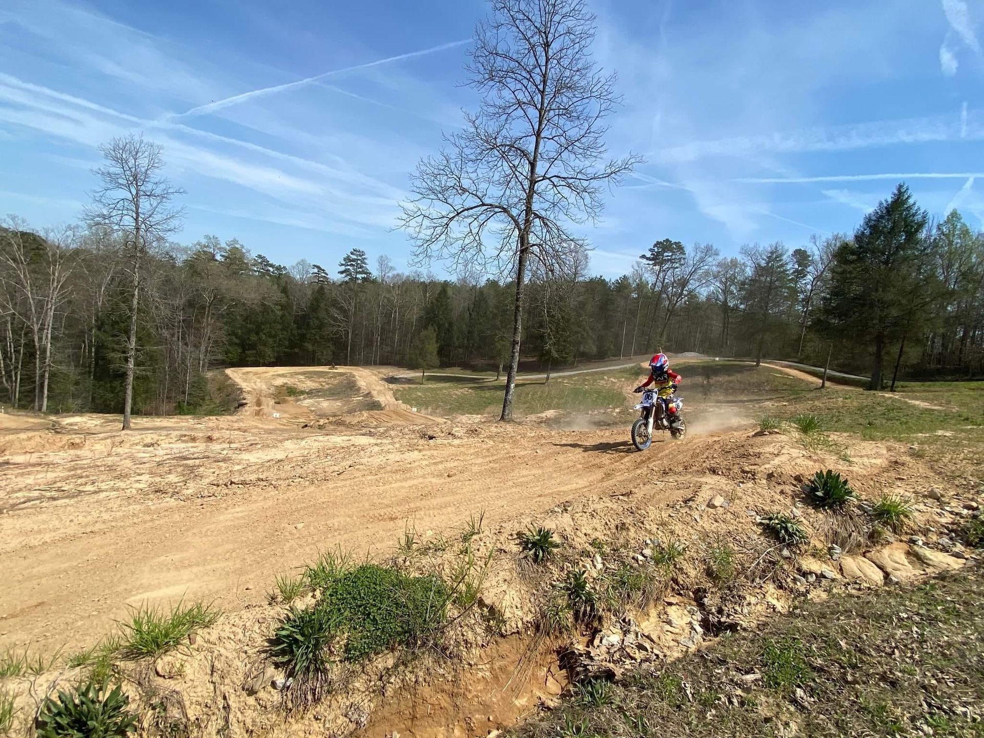 A motocross rider speeds along a dirt track on a sunny day with trees in the background.