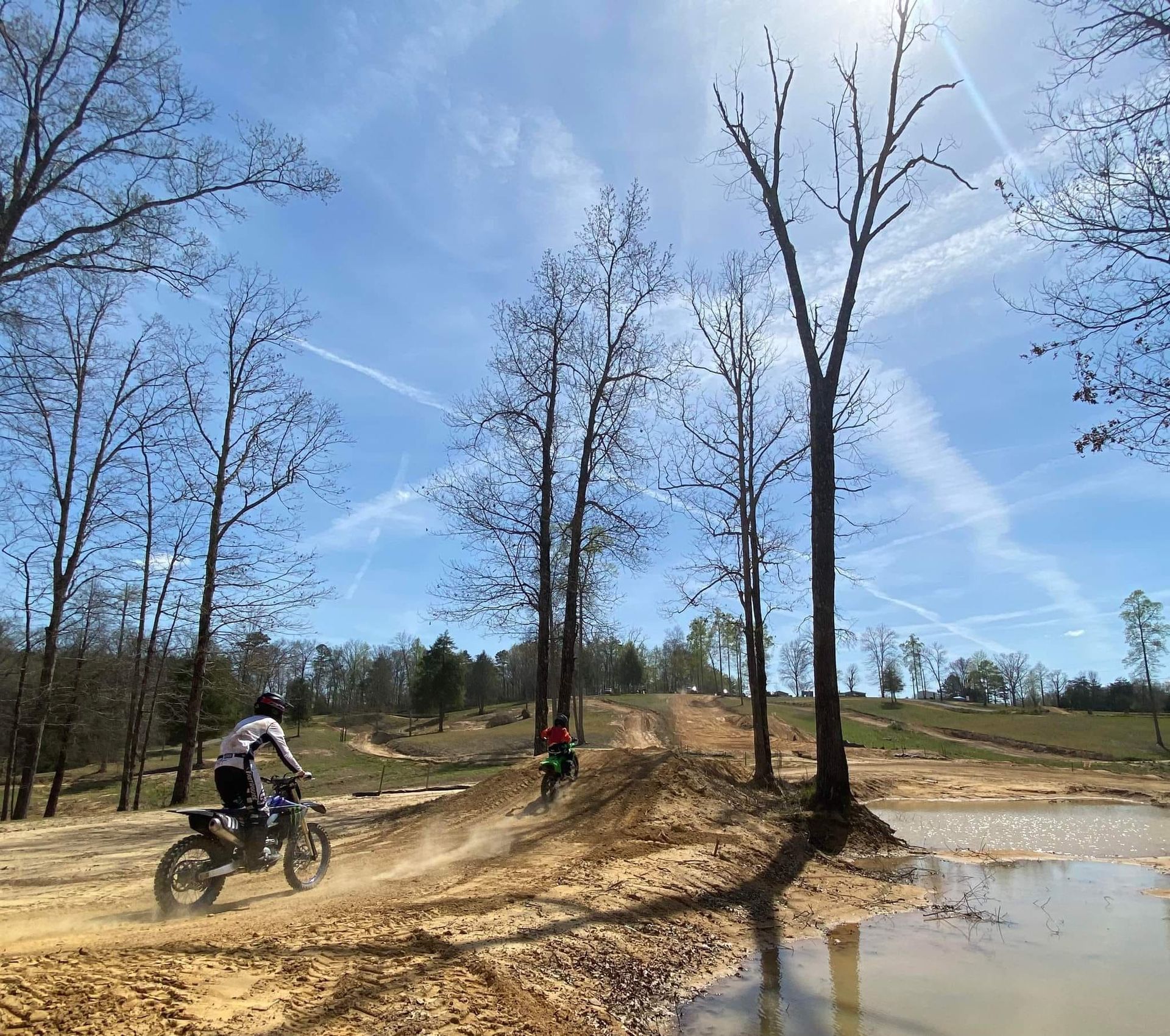 Two dirt bikers ride on a sandy trail under a clear blue sky, passing a small pond near a line of bare trees.
