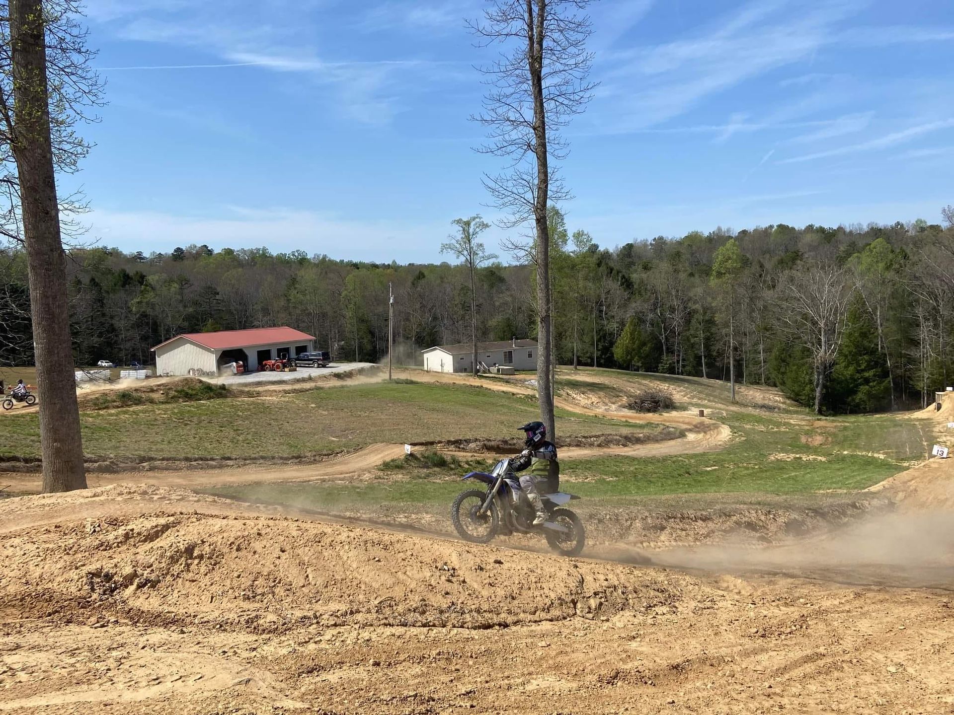 A motorcyclist rides across a dirt track under a bright blue sky, with rural buildings and a forest in the background.