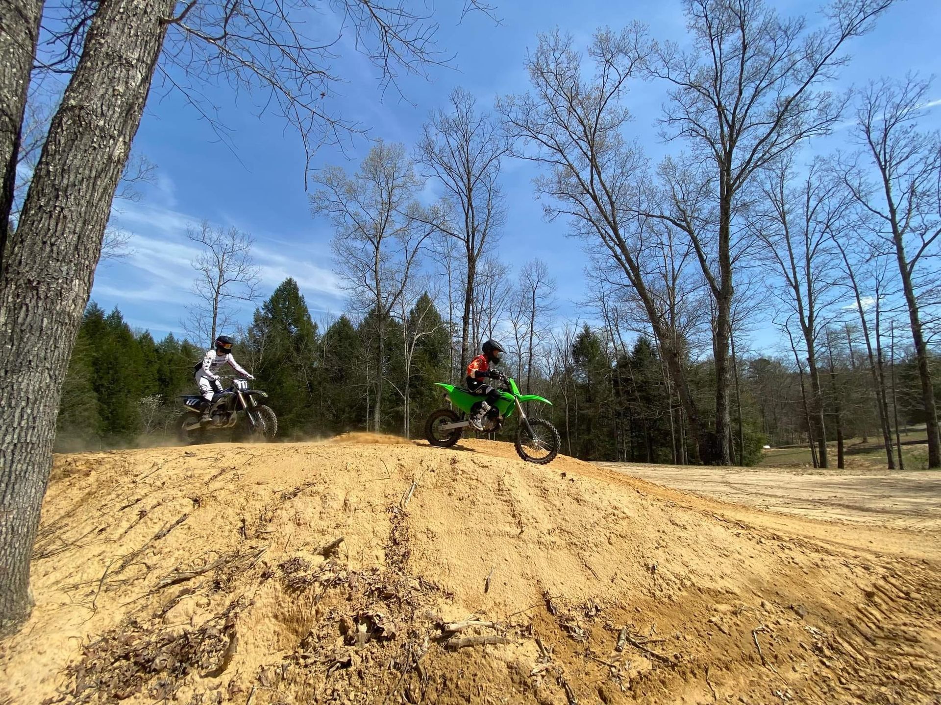 Two motorcyclists ride dirt bikes over a large sand hill in a wooded area on a sunny day.