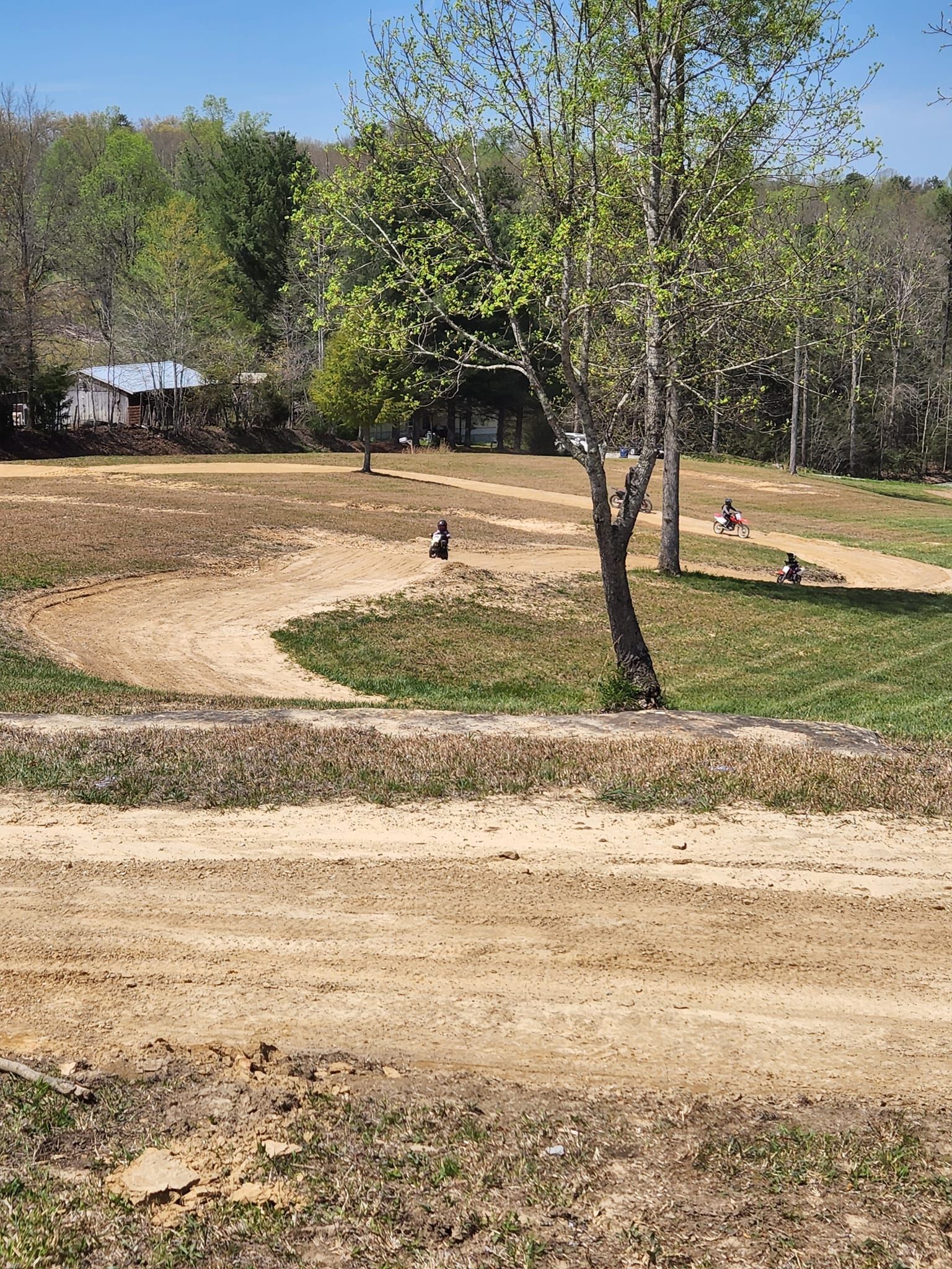 Dirt bike track with several riders practicing on a course featuring hills and curves on a sunny day.