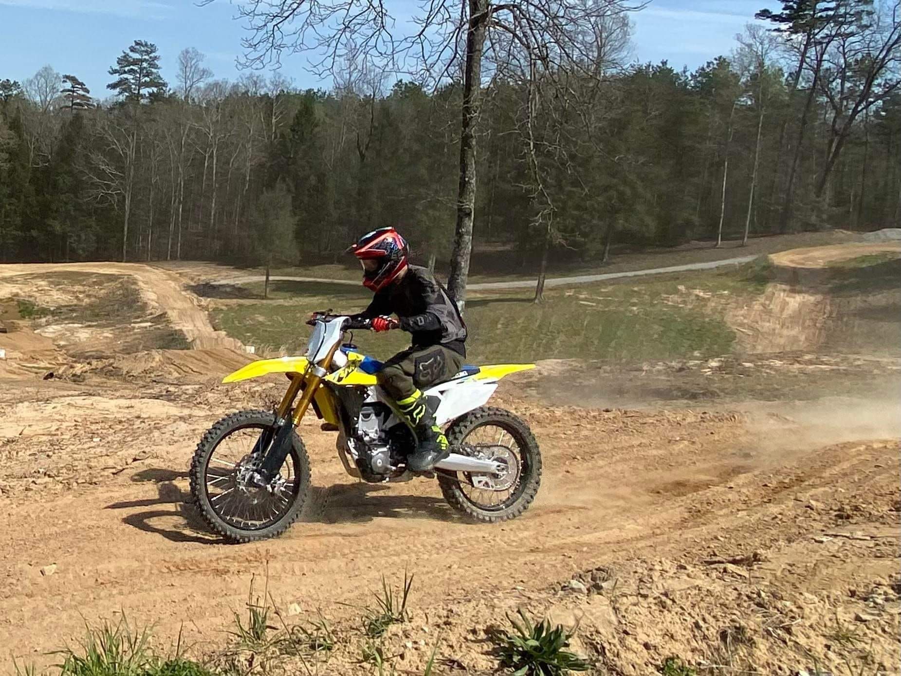 A rider on a yellow dirt bike speeding across a sunny, dirt-covered outdoor track with trees in the background.