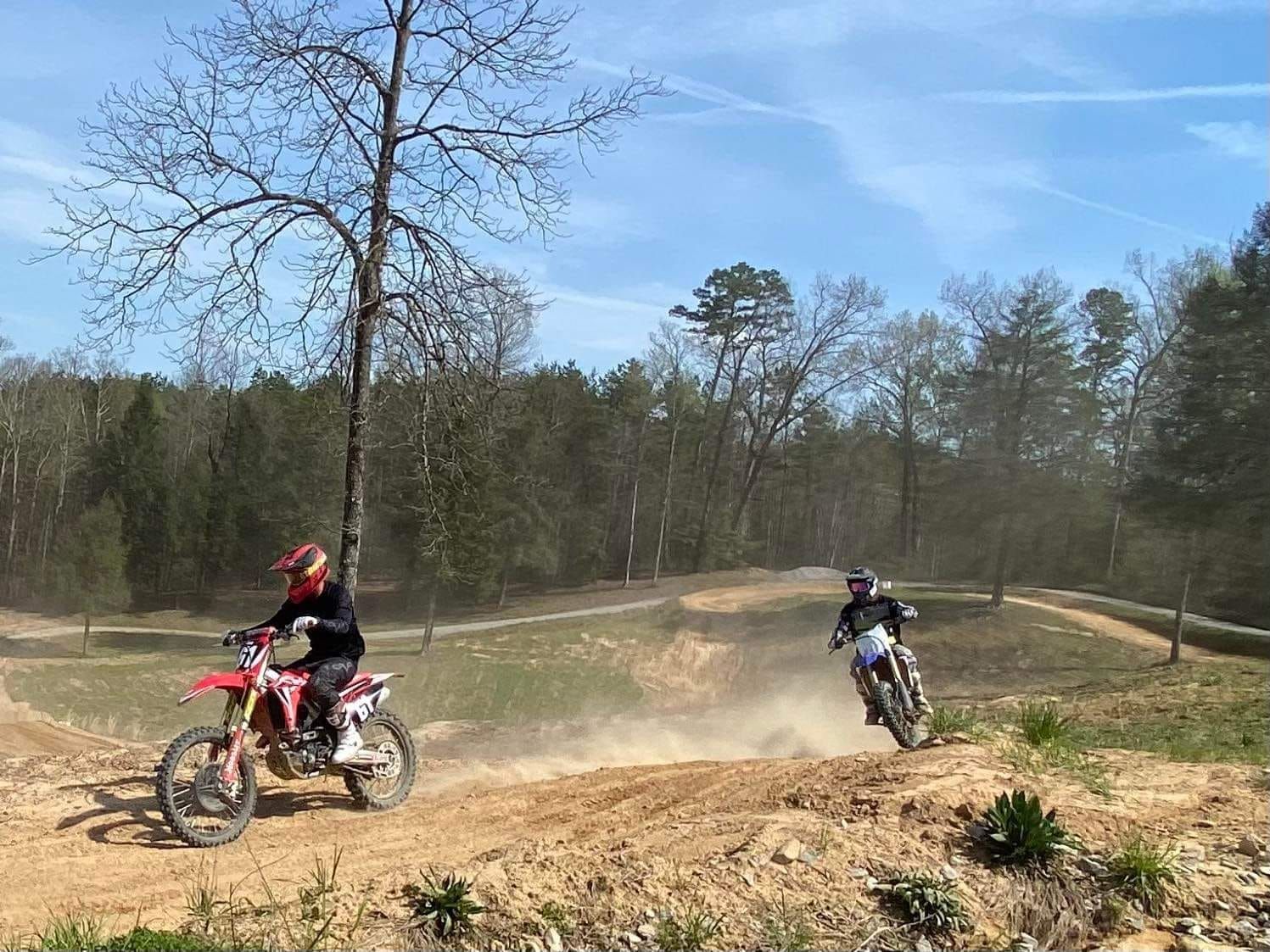 Two people ride dirt bikes on a dirt track in a wooded area on a sunny day.