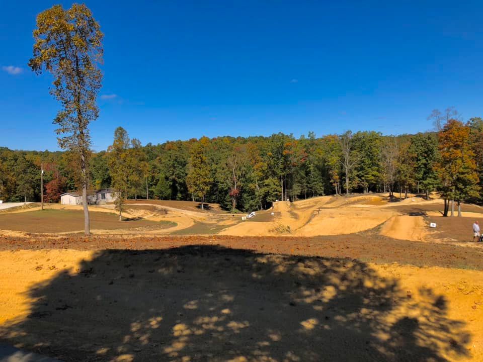 An outdoor motocross dirt track with jumps, surrounded by a dense forest under a clear blue sky.