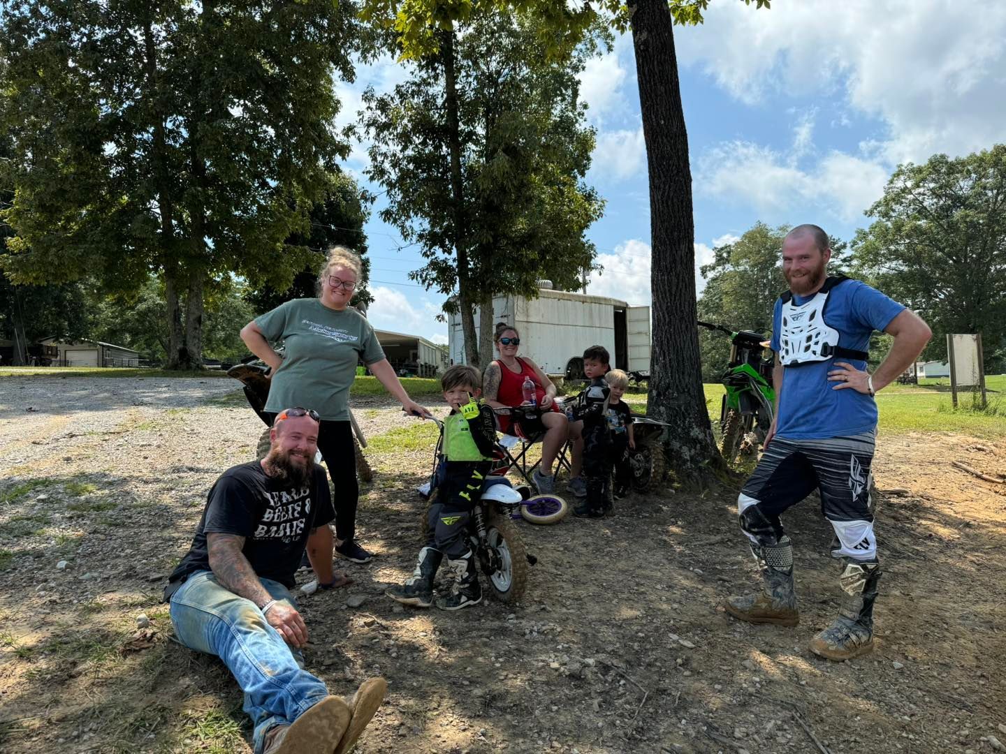 A group of people gathers outdoors in a shaded area with dirt bikes, some standing and others sitting near a large tree.