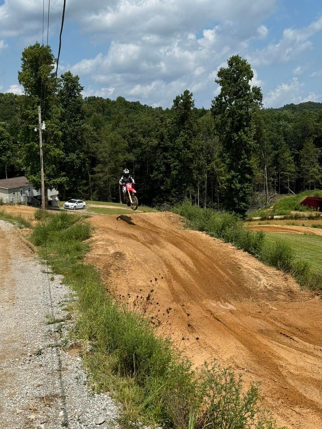 A rider on a red dirt bike jumps over a brown earthen mound on an outdoor motocross track with trees in the background.