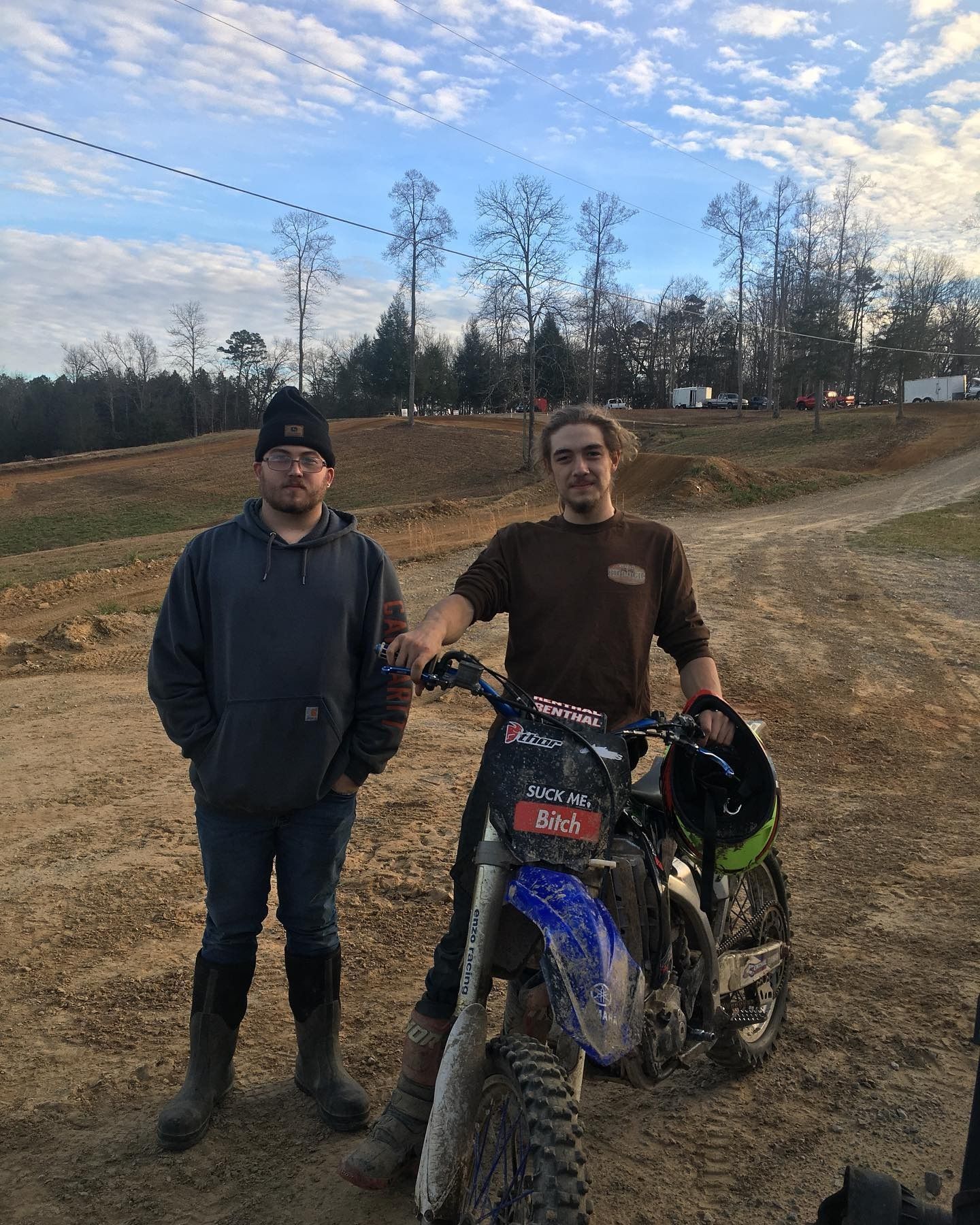 Two people pose with a blue dirt bike on a dirt track during the day under a blue sky with scattered clouds.