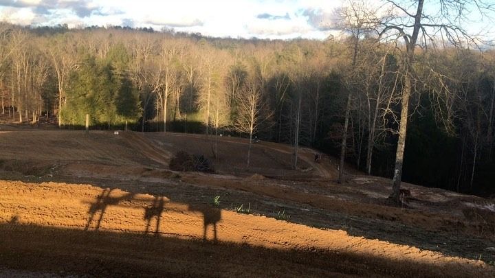A cleared, muddy construction site at the edge of a forest, with long shadows cast across the foreground.