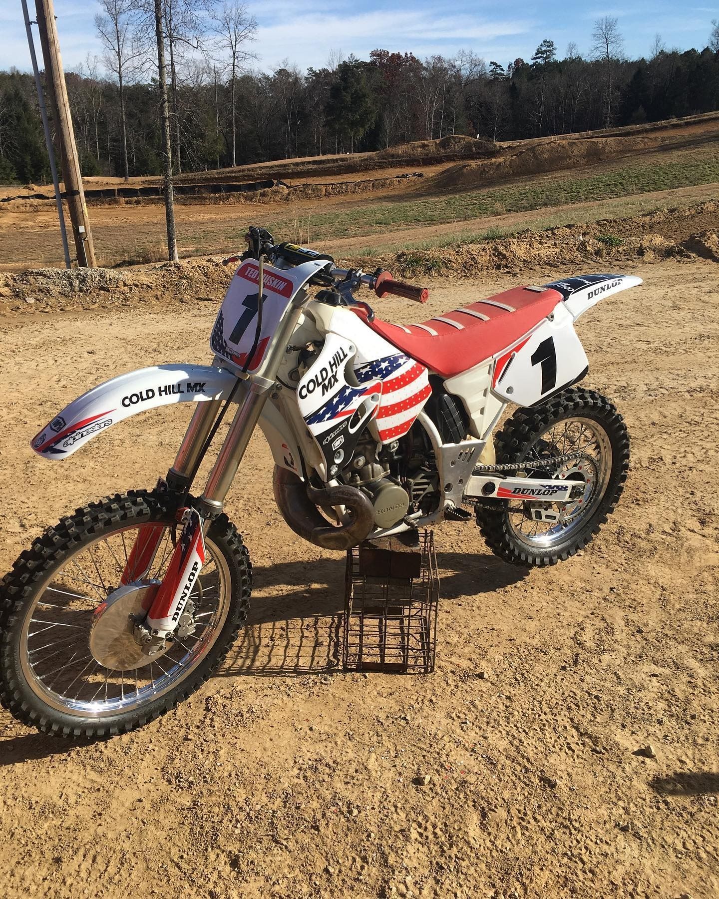 A white and red dirt bike with an American flag graphic on a metal stand in an outdoor, dirt-filled field.