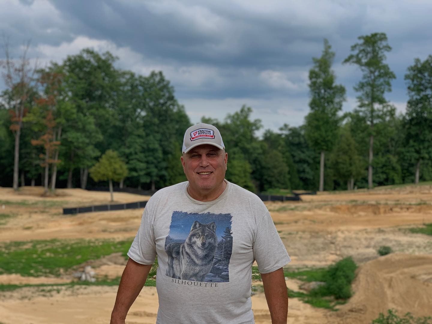 A person wearing a wolf graphic t-shirt and a baseball cap stands outdoors in front of a dirt construction site.