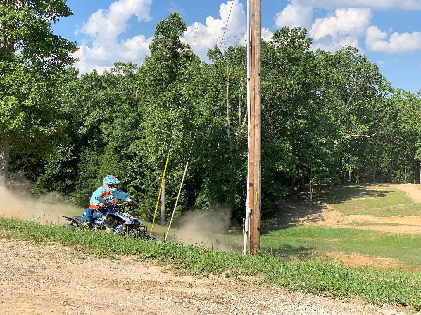 A person on an ATV speeds through a dirt track, kicking up dust against a backdrop of trees and a utility pole.