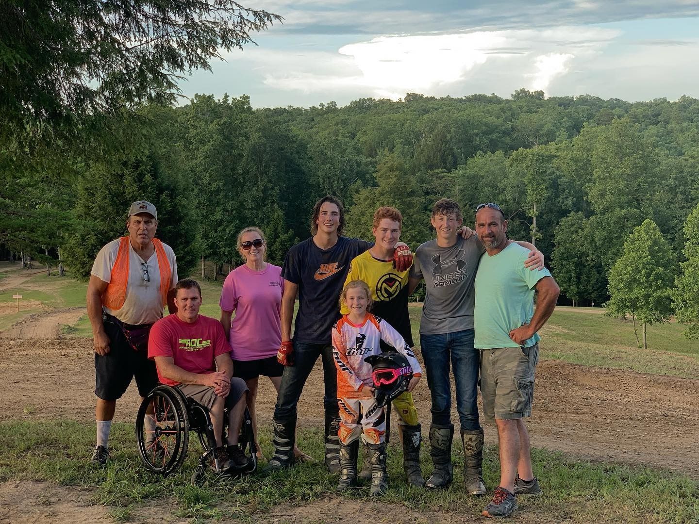 A group of eight people stand and sit together on a dirt trail in front of a forest, smiling for a photo.