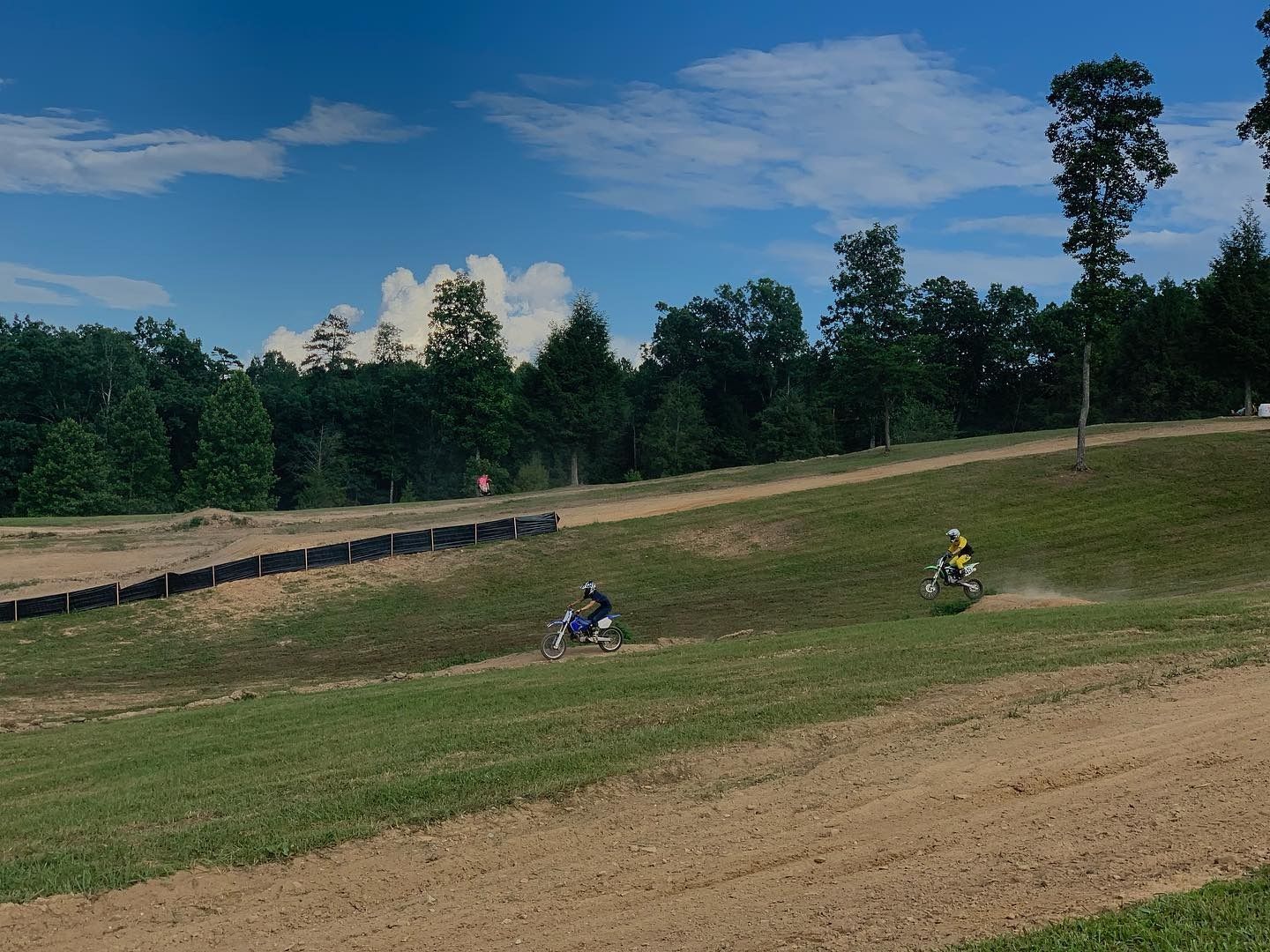 Two motorcyclists riding on a grassy dirt track under a bright blue sky with scattered clouds.