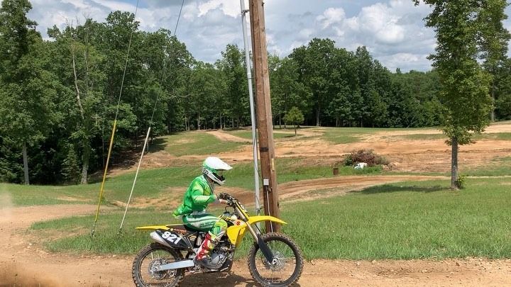 A rider in green gear on a yellow dirt bike riding on a dirt track near a utility pole and trees.