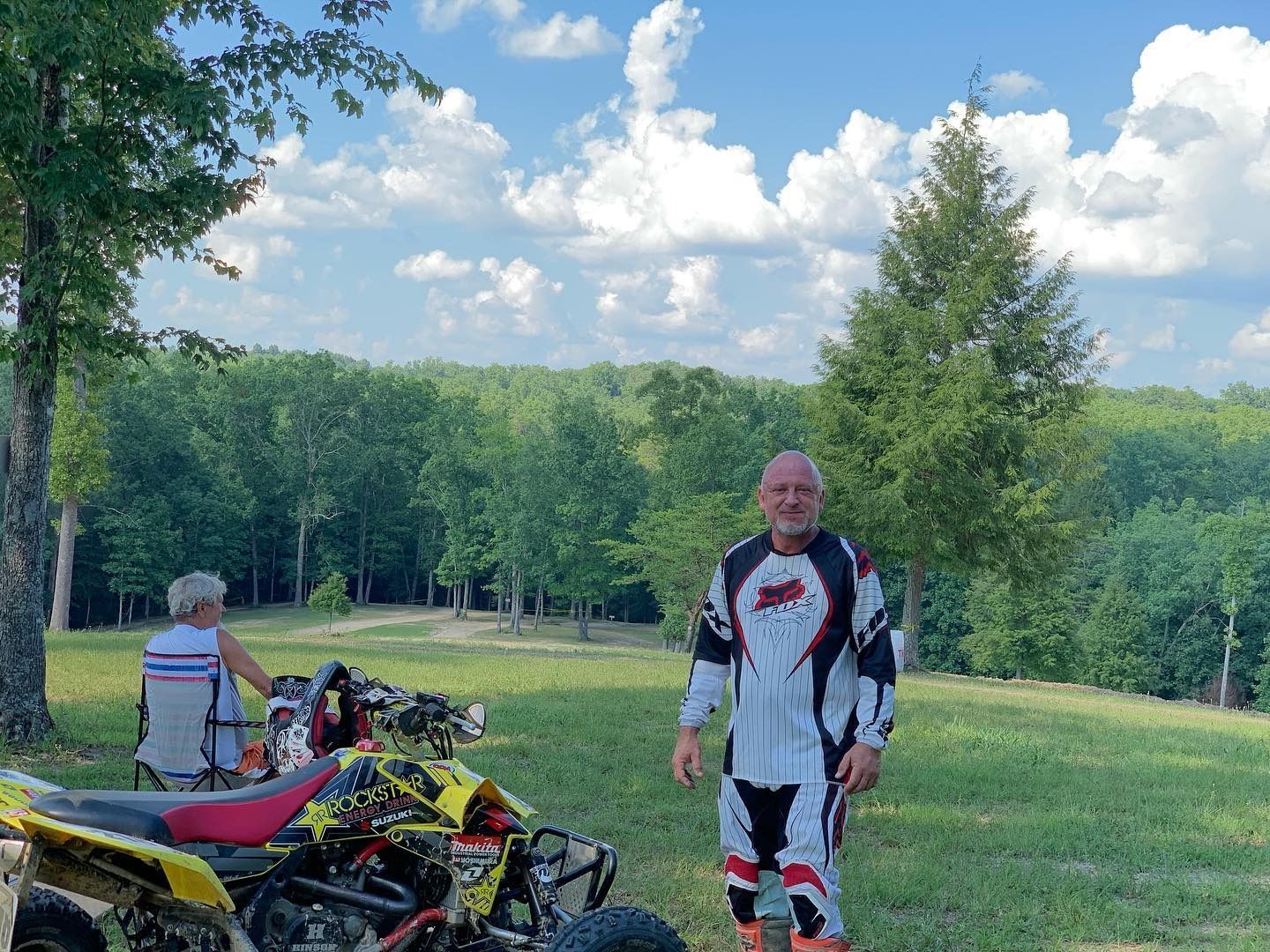 A person in motocross gear stands by an ATV on a grassy hill overlooking a forest under a blue sky with clouds.