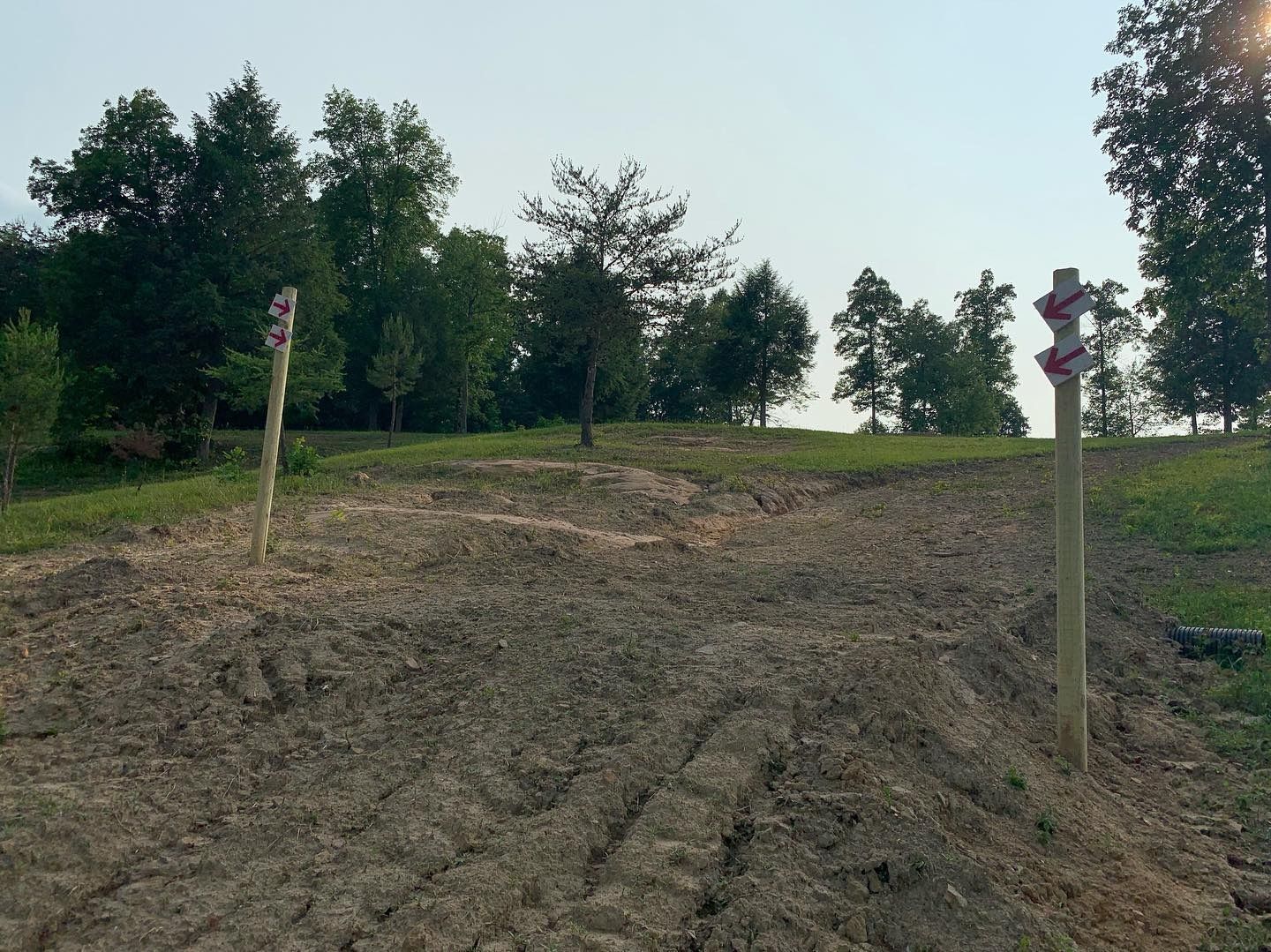 Two posts with red trail markers stand on a path through a freshly cleared dirt area leading to trees and a grassy hill.