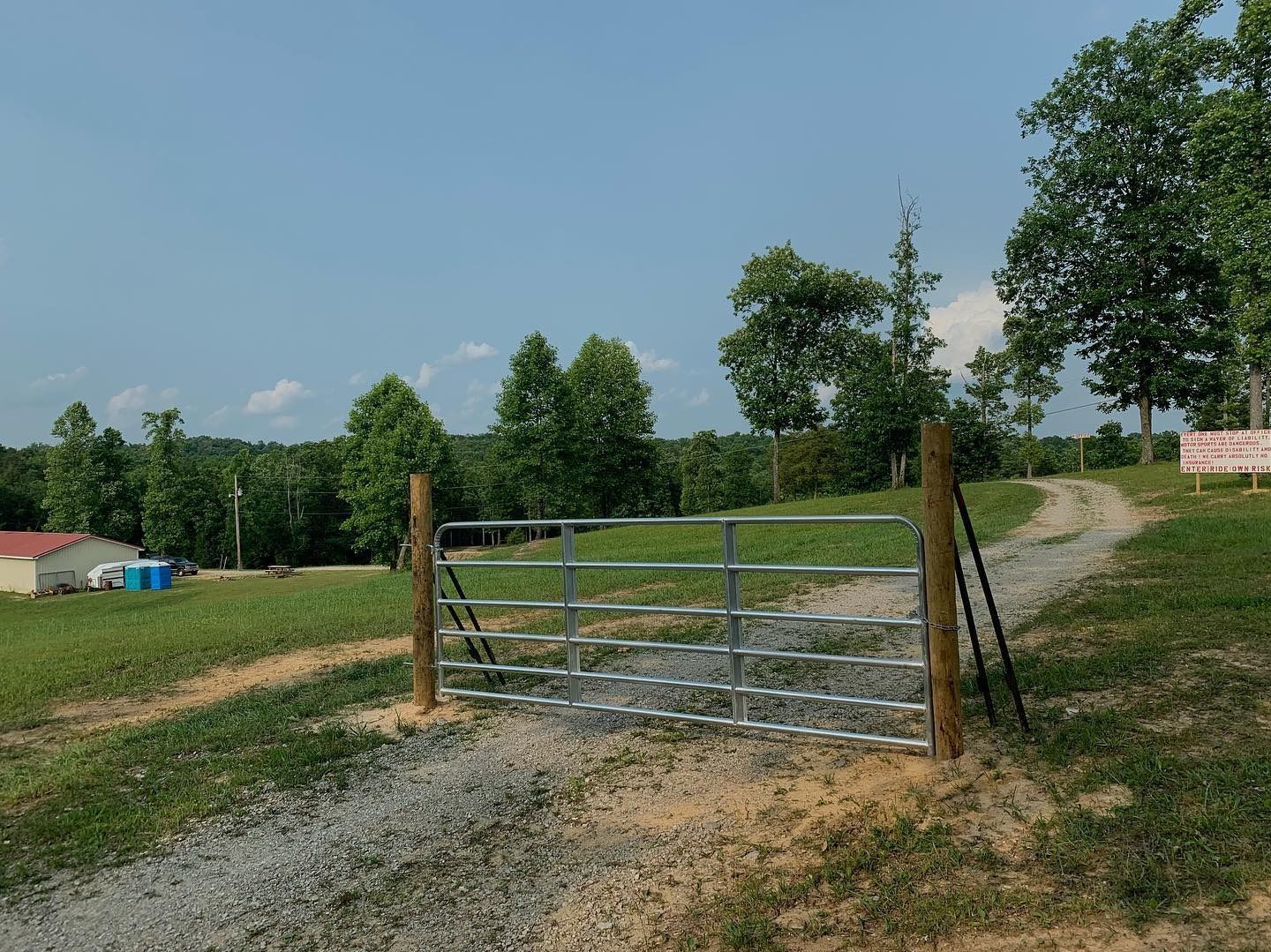 A metal gate stands at the entrance of a gravel driveway on a grassy, rolling field under a clear blue sky.