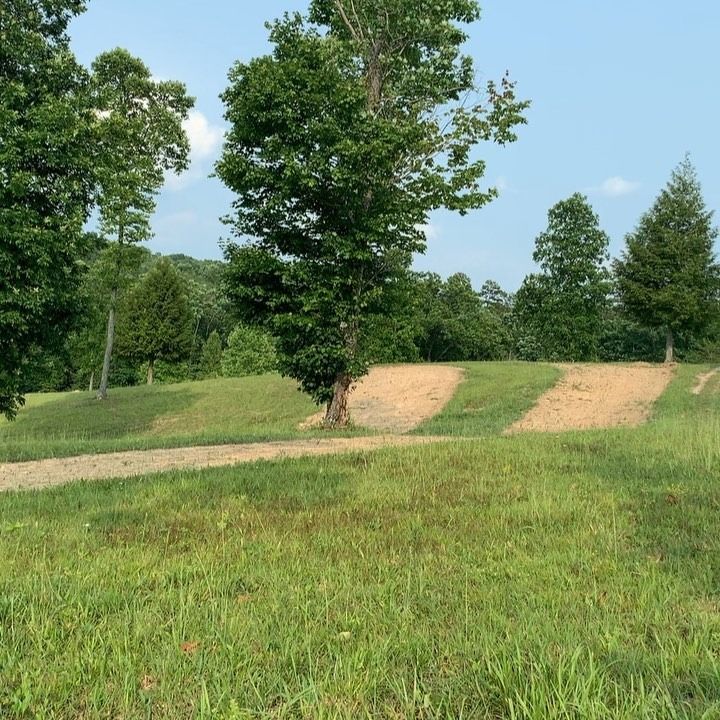A landscape view of a green grassy field with trees and bare, soil-covered strips running across a gentle hill.
