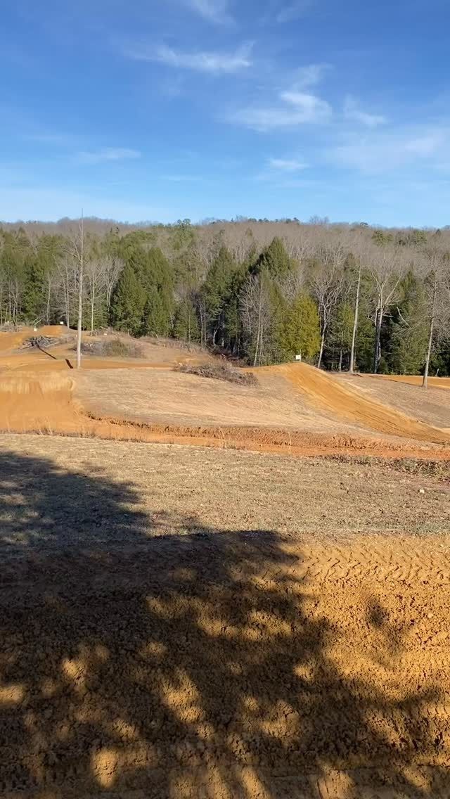 A dirt bike track with earthen jumps and mounds sits under a clear blue sky, framed by a line of trees in the background.