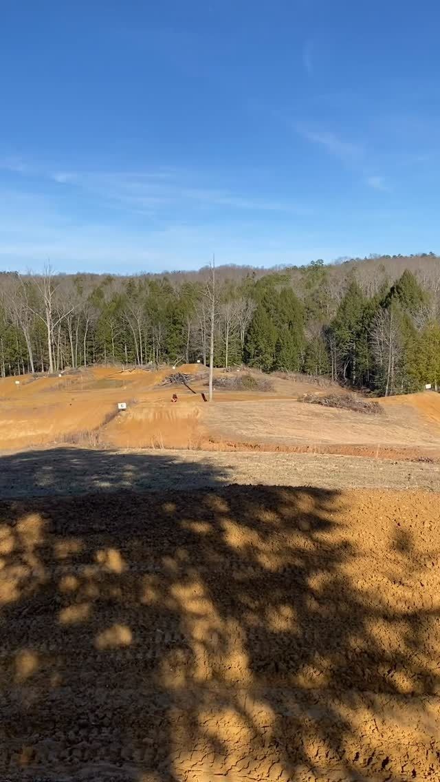 A dirt construction or riding area with mounds of earth in front of a forest line under a clear blue sky.