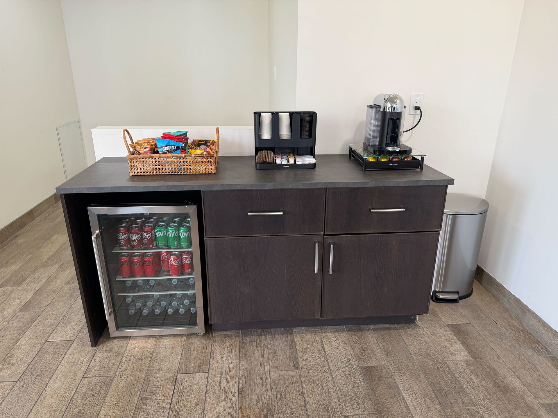 Coffee station with a mini-fridge, coffee maker, and snacks on a dark cabinet against a white wall | Euro Auto Repair