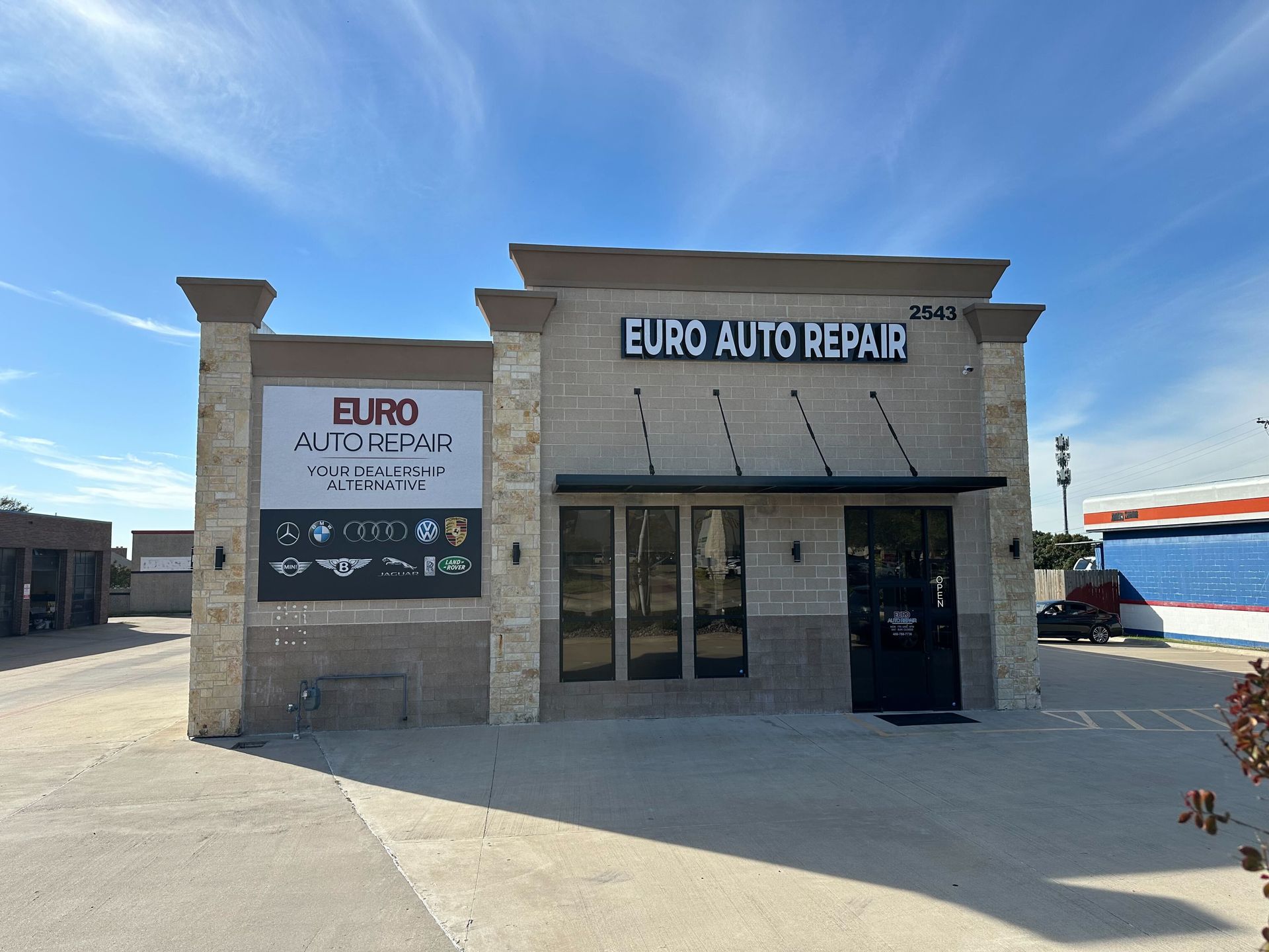Euro Auto Repair storefront under a blue sky. Light-colored brick facade, black doors and window trim, a sign with the business name | Euro Auto Repair