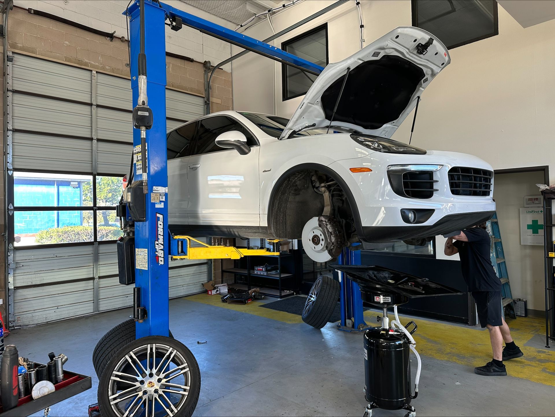A white Porsche SUV on a lift in a garage with a mechanic working on it | Euro Auto Repair