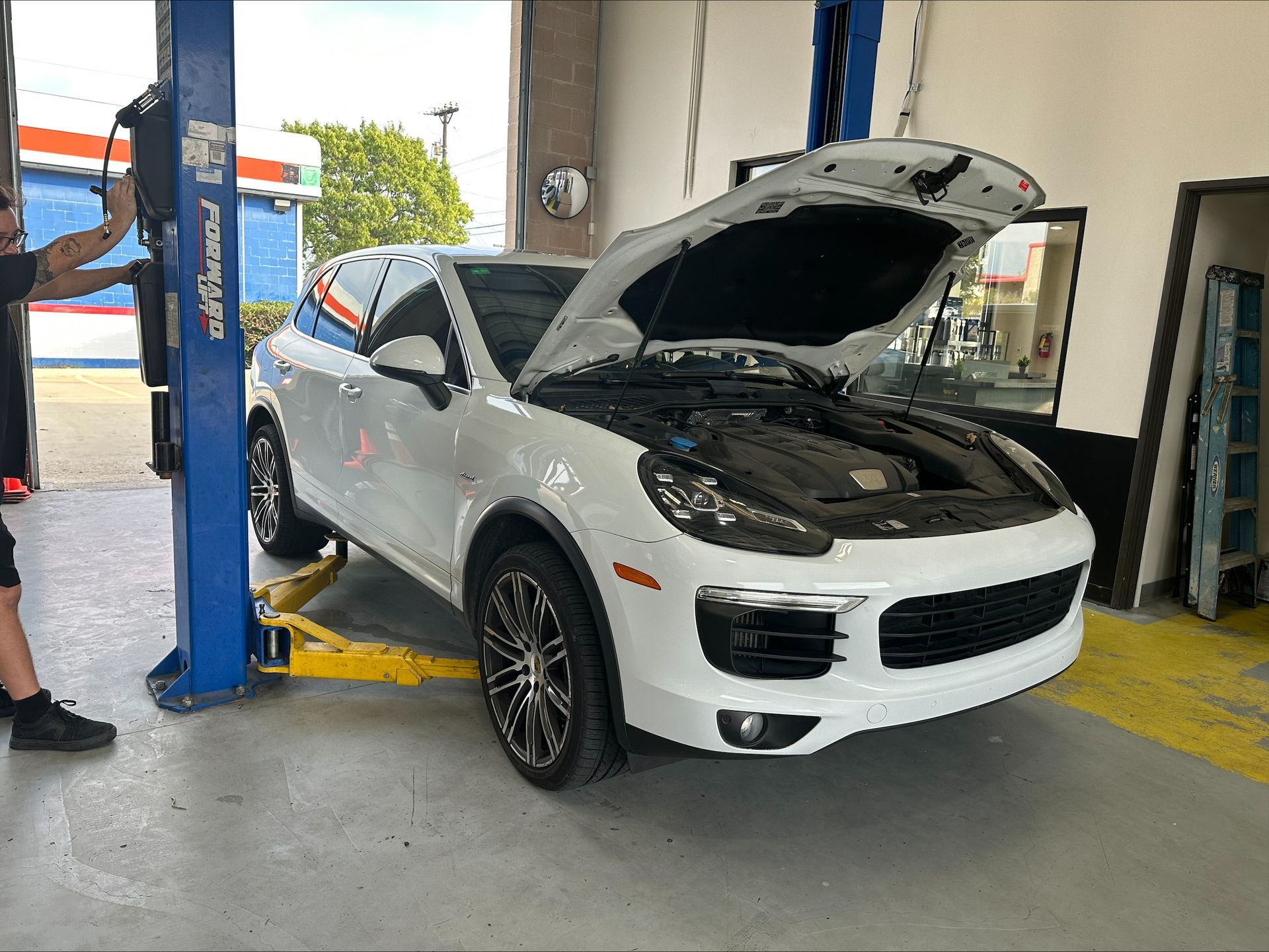 White Porsche Cayenne on a lift in a repair shop with its hood open; a mechanic is visible | Euro Auto Repair
