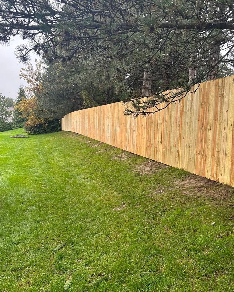 A long, newly installed wooden privacy fence runs along a sloped, grassy lawn beneath mature evergreen trees.