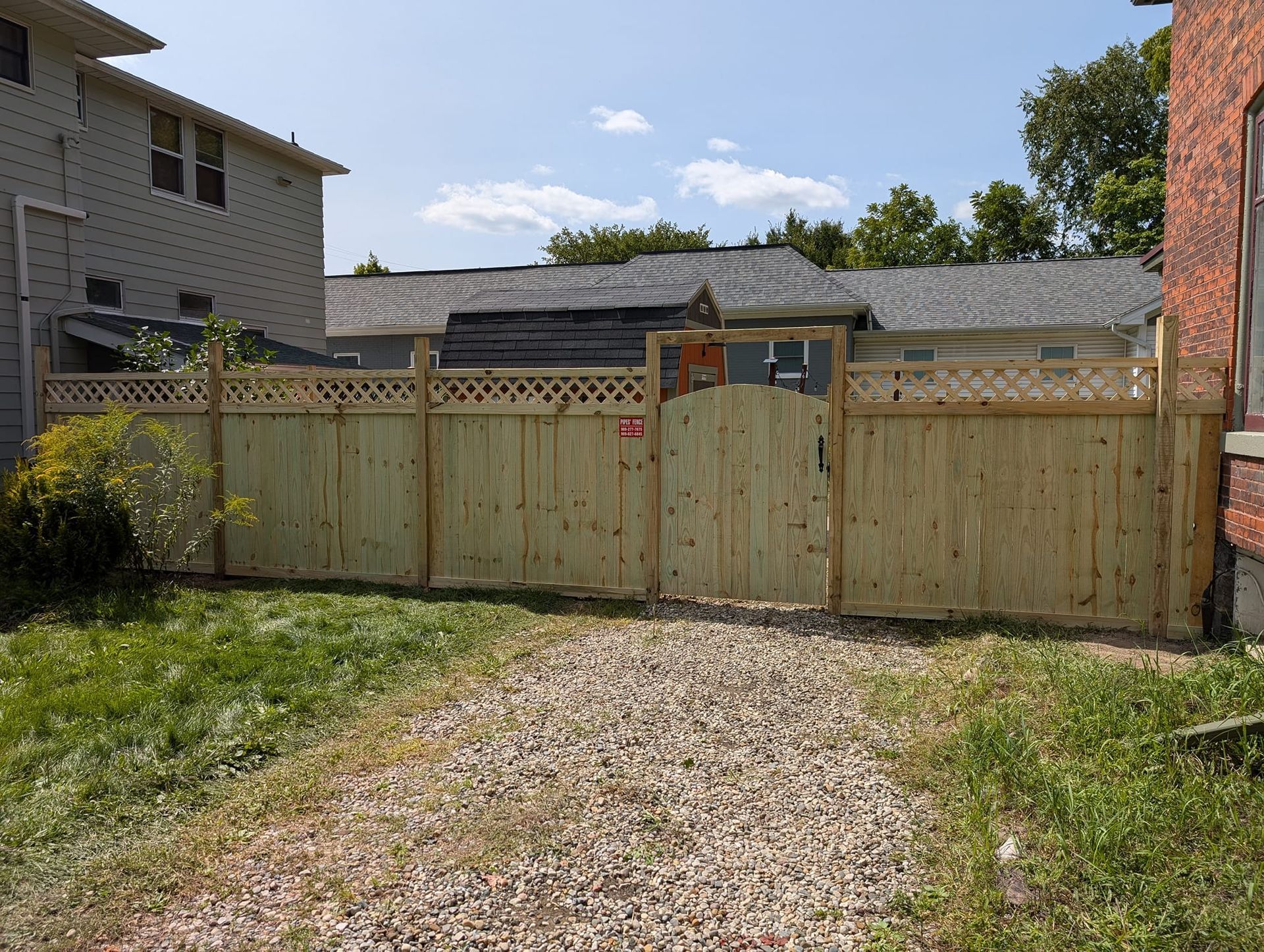 A new wooden privacy fence with a lattice top and a central arched gate, situated on a gravel driveway between two houses.