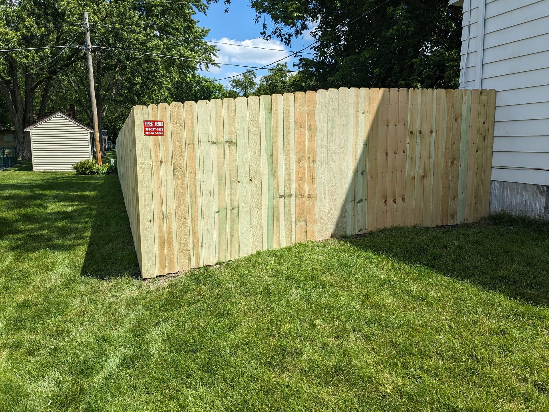 A newly installed light-wood privacy fence standing on a green lawn beside a white house under a sunny blue sky.
