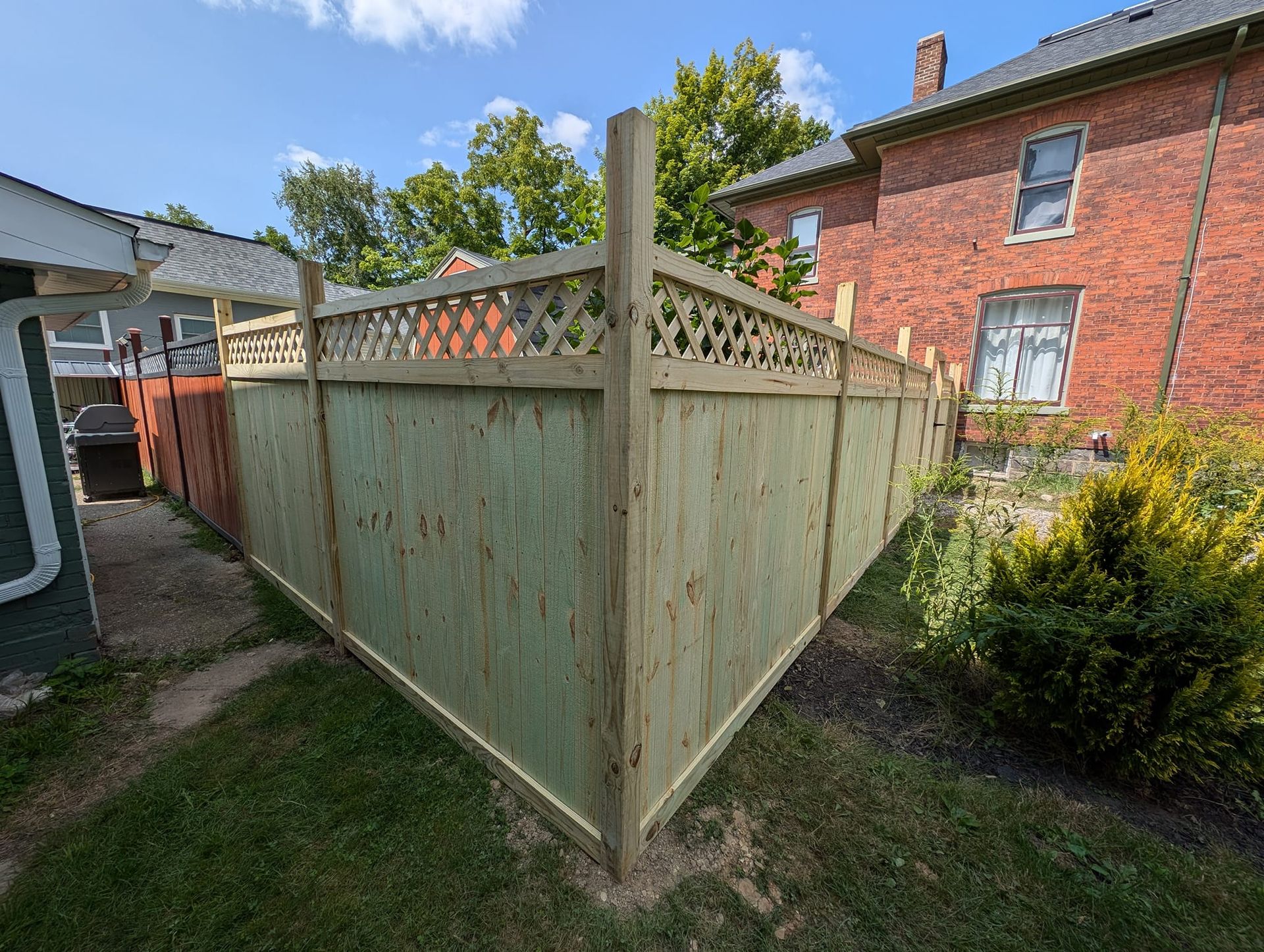 A new, light-colored wooden privacy fence with a decorative lattice top forming a corner in a grassy backyard.