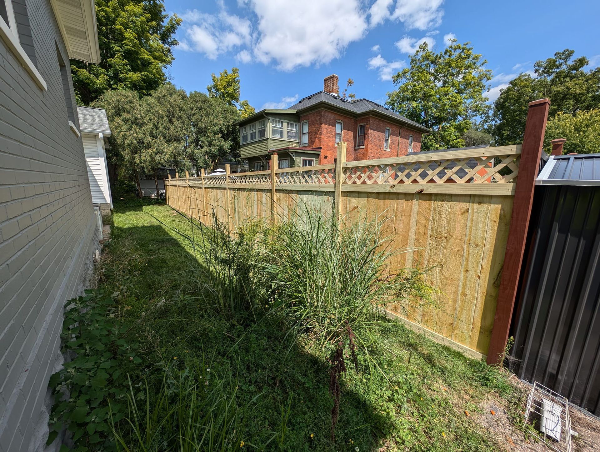 A side yard shows a new wooden fence with lattice topping, running alongside a grey house and overgrown weeds.