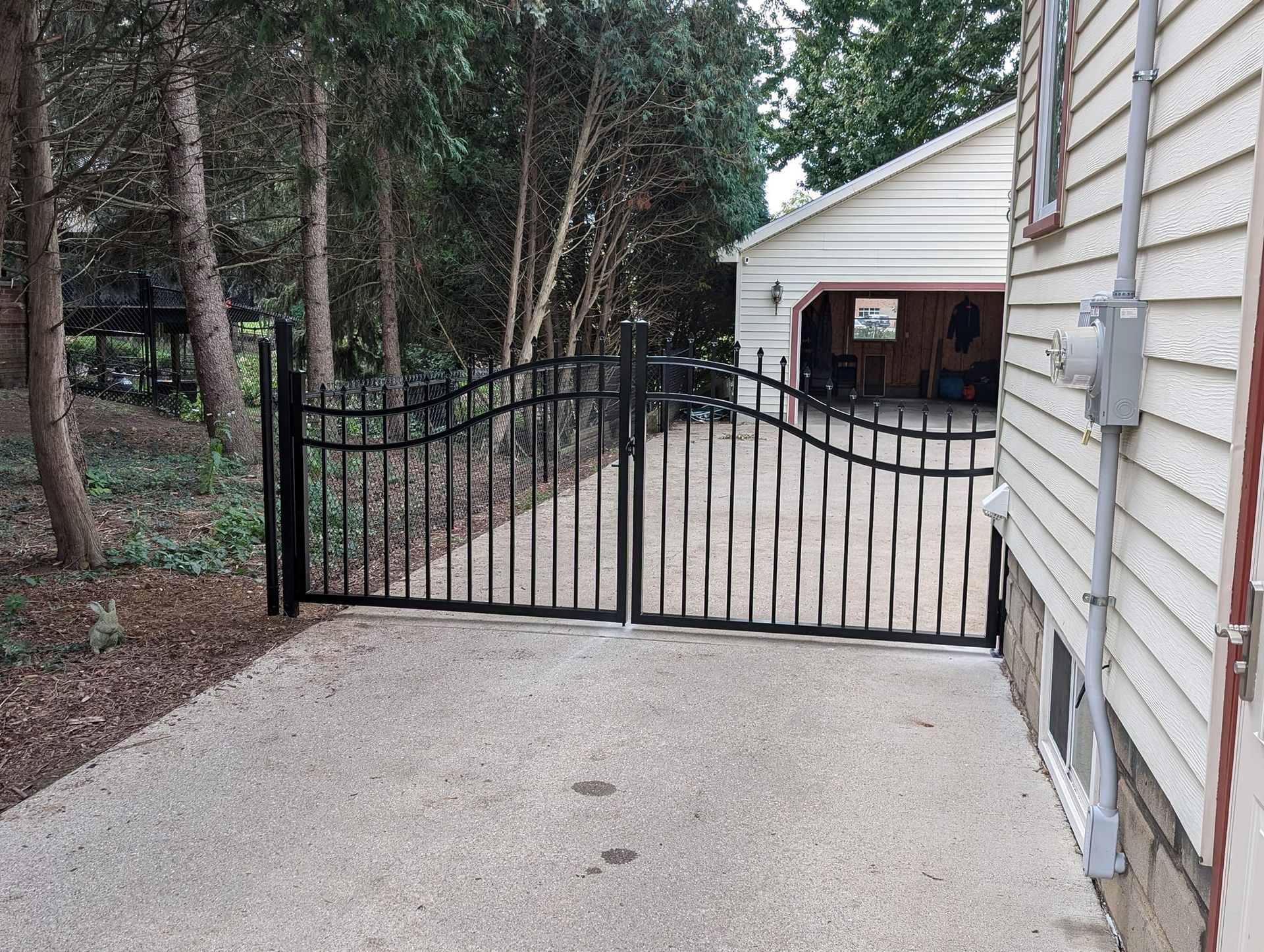 A black arched metal driveway gate installed between a wooded area and the side of a white house with a garage behind.