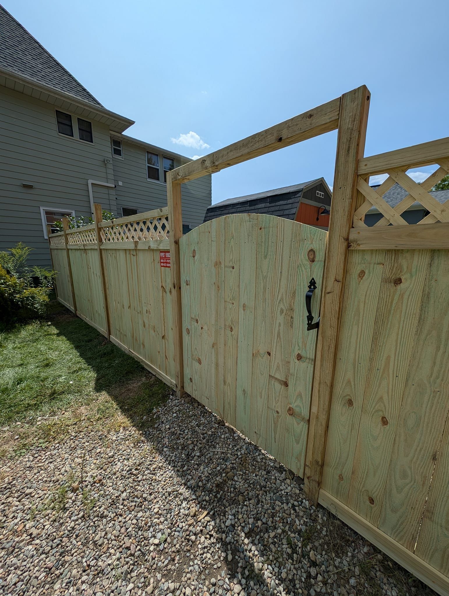 A light-wood privacy fence and arched gate with a lattice topper, set on a gravel path beside a grey house.