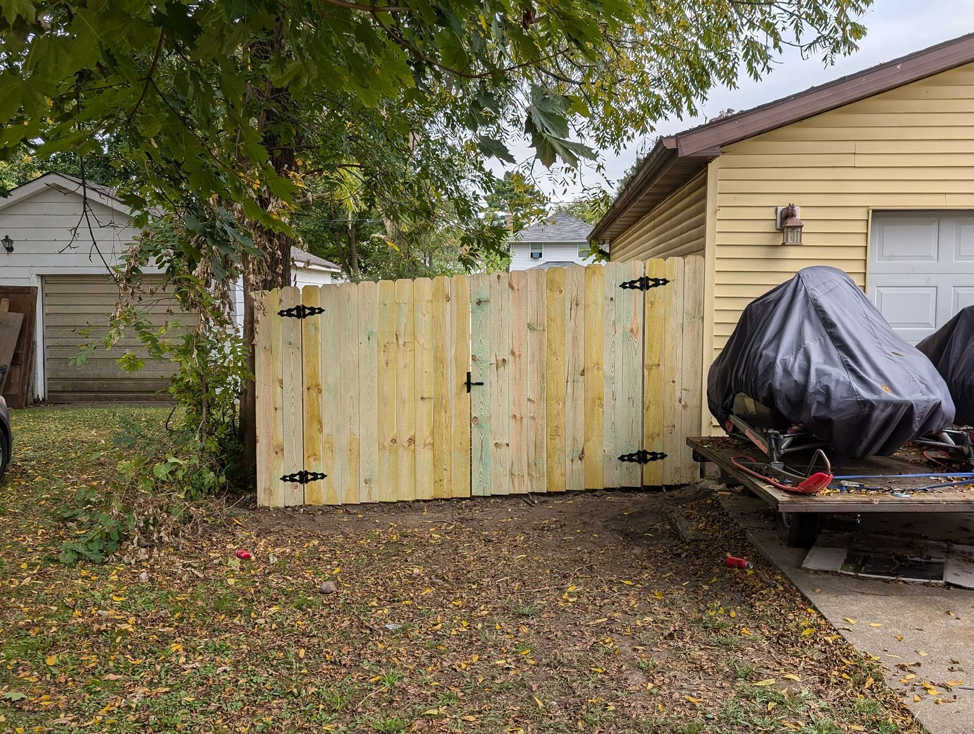 A new wooden double gate stands between a garage and a tree-filled yard, with a covered vehicle on a trailer nearby.