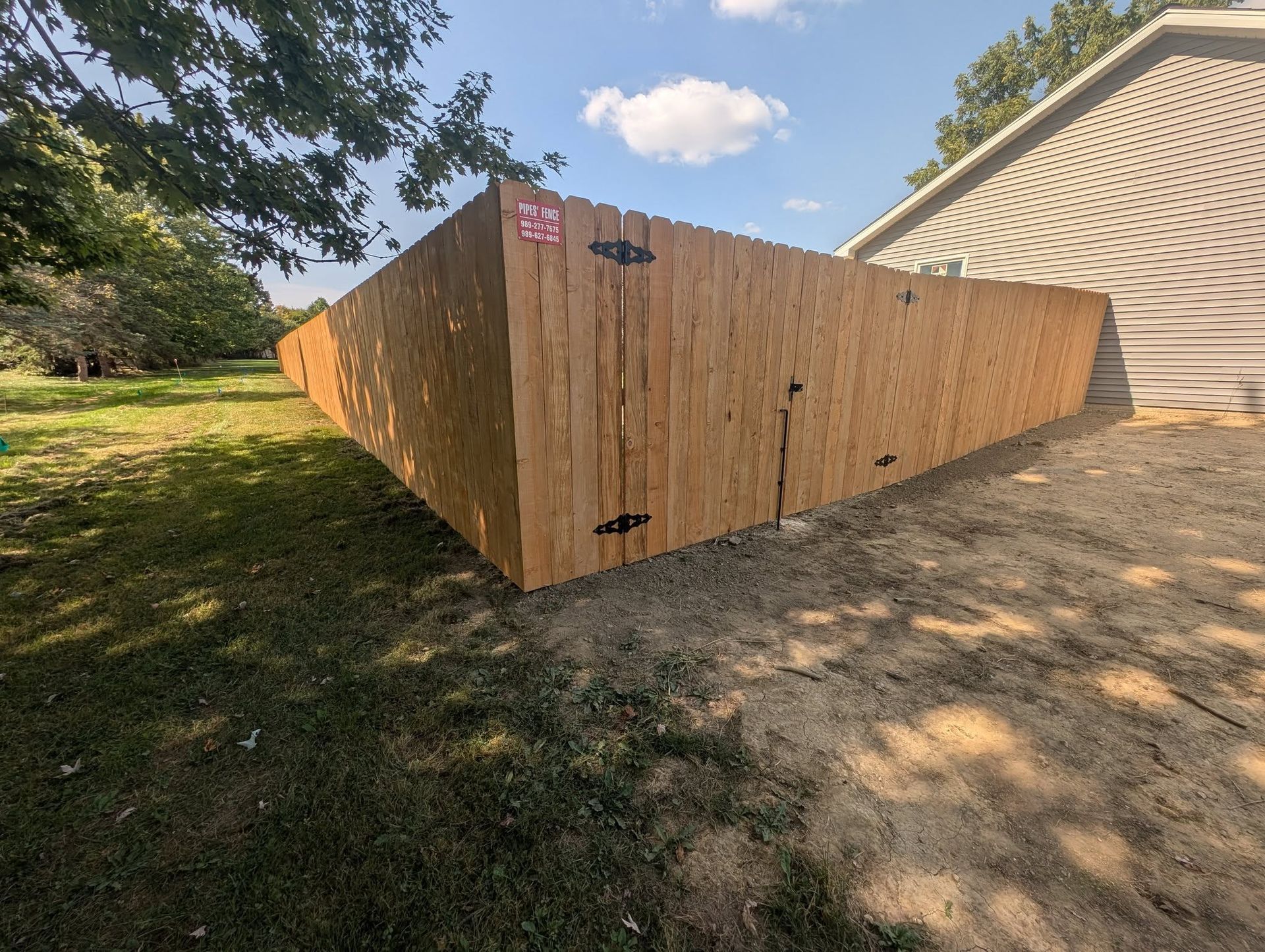 A wooden privacy fence extends from the corner of a house exterior into a grassy yard under a blue sky.