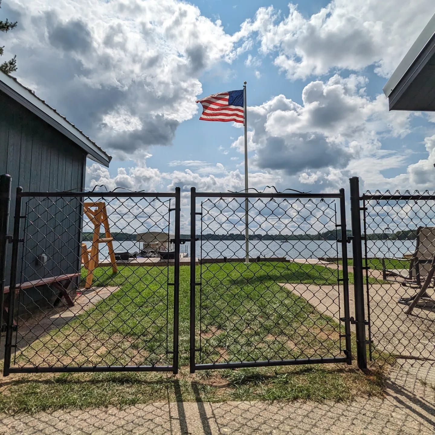 An American flag flies above a chain-link gate leading to a lakeside yard under a bright, cloudy sky.