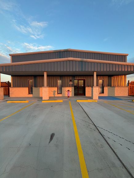 Building with a unique zig-zag roof, person standing in front with arms raised, clear sky, parking lot.