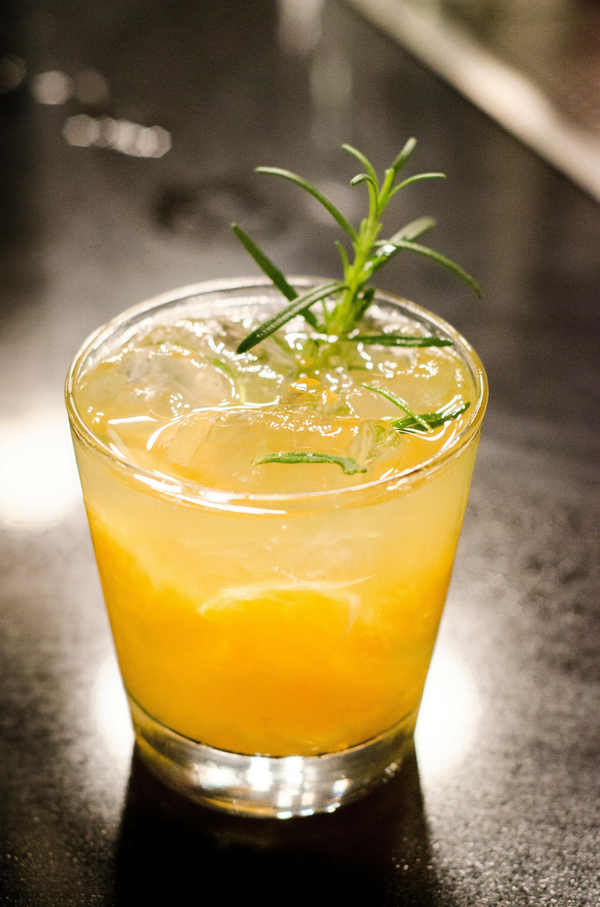 Yellow cocktail in a glass with ice and a sprig of rosemary.