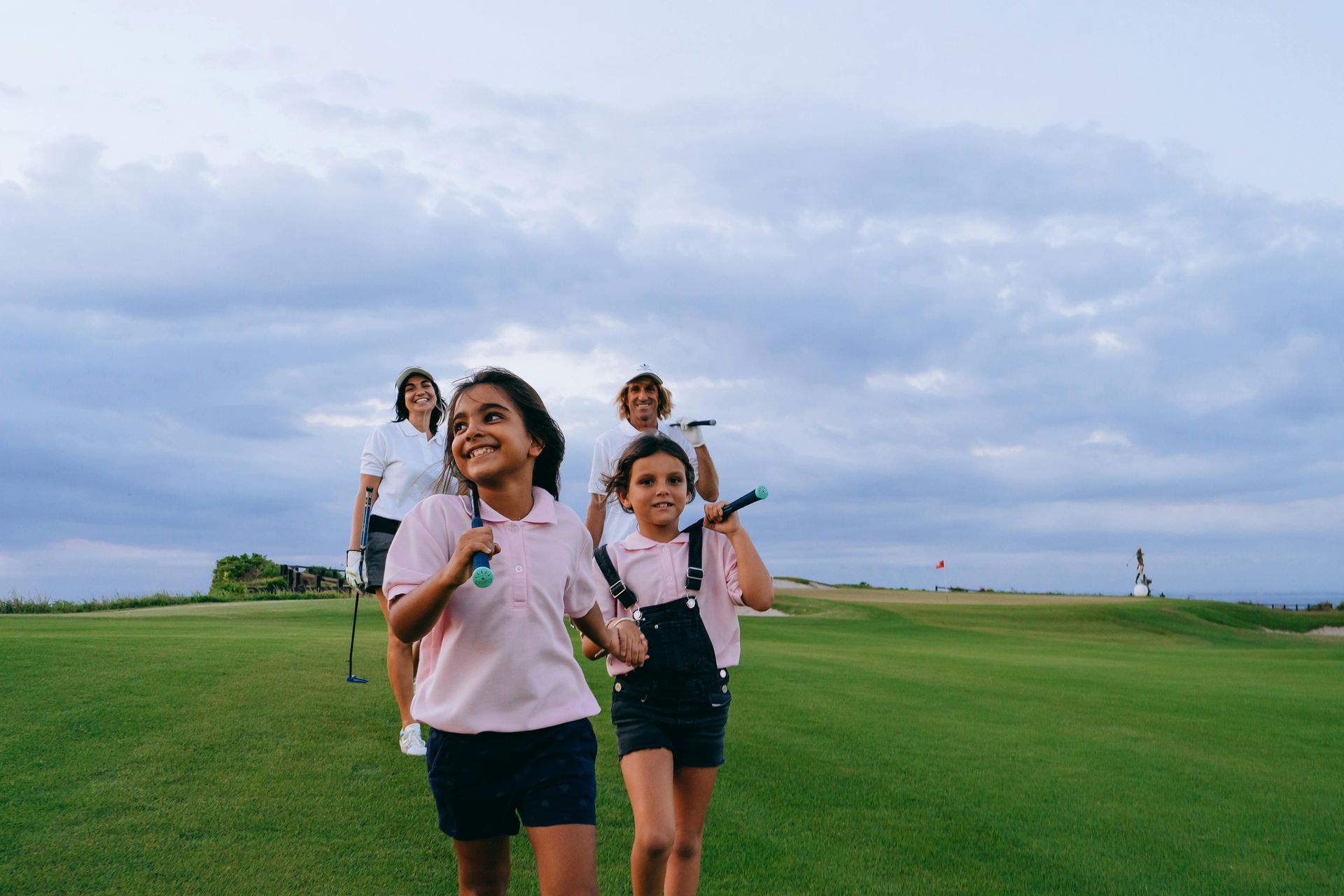 Two girls in pink shirts and overalls walk on a golf course, holding clubs. Two adults follow behind.