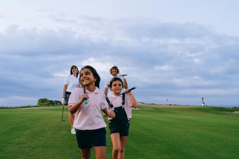 Two girls in pink shirts and overalls walk on a golf course, holding clubs. Two adults follow behind.