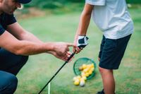 Man teaching a child how to grip a golf club on a green field.