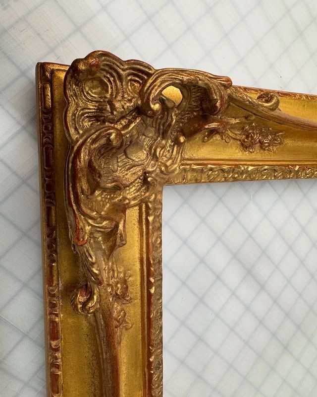 Ornate, gilded gold wooden picture frame corner with intricate scrollwork, resting on a grid-patterned surface.