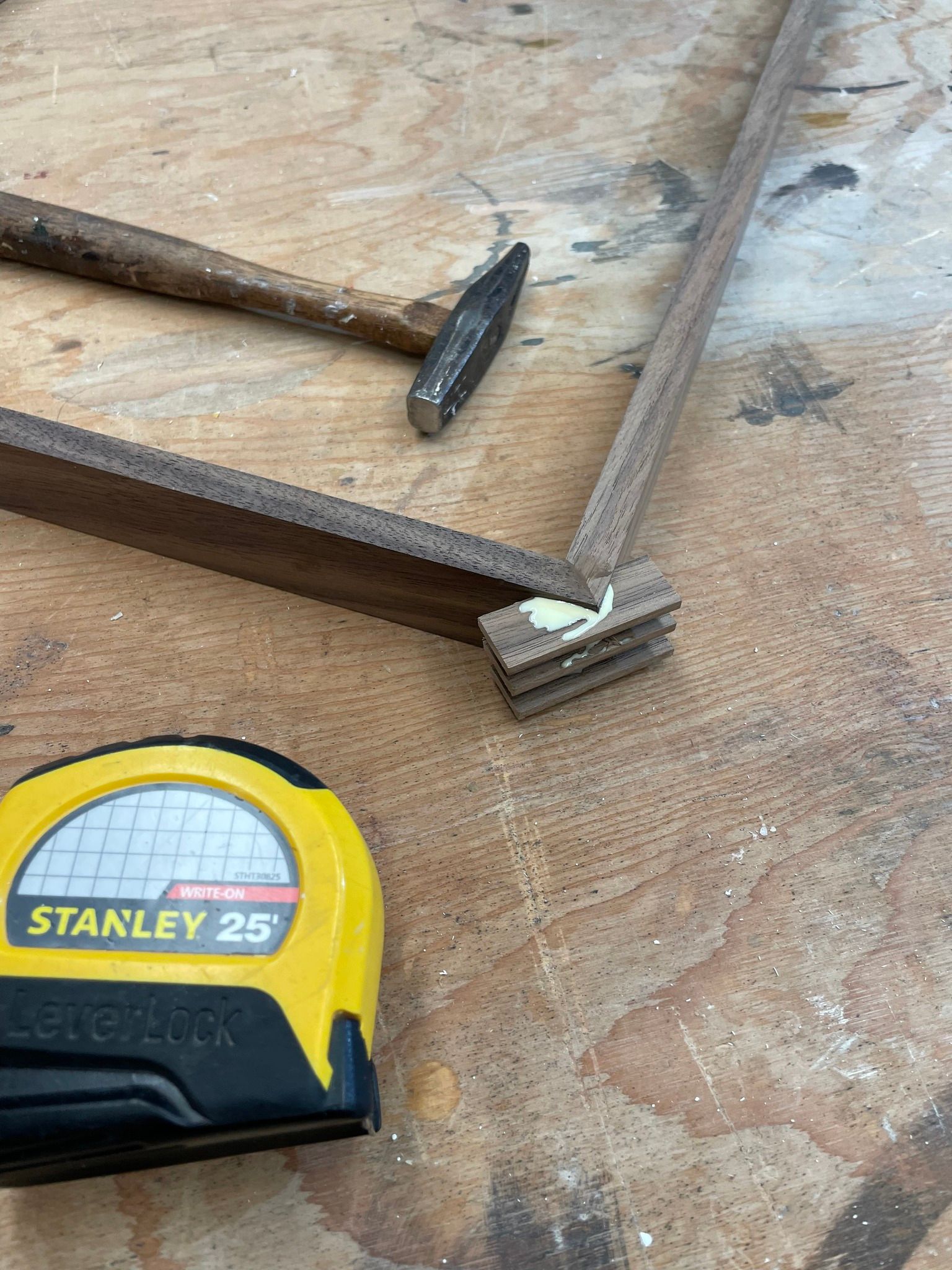 A Stanley tape measure, a small hammer, and two wooden frame pieces joined at a corner with adhesive on a wooden surface.